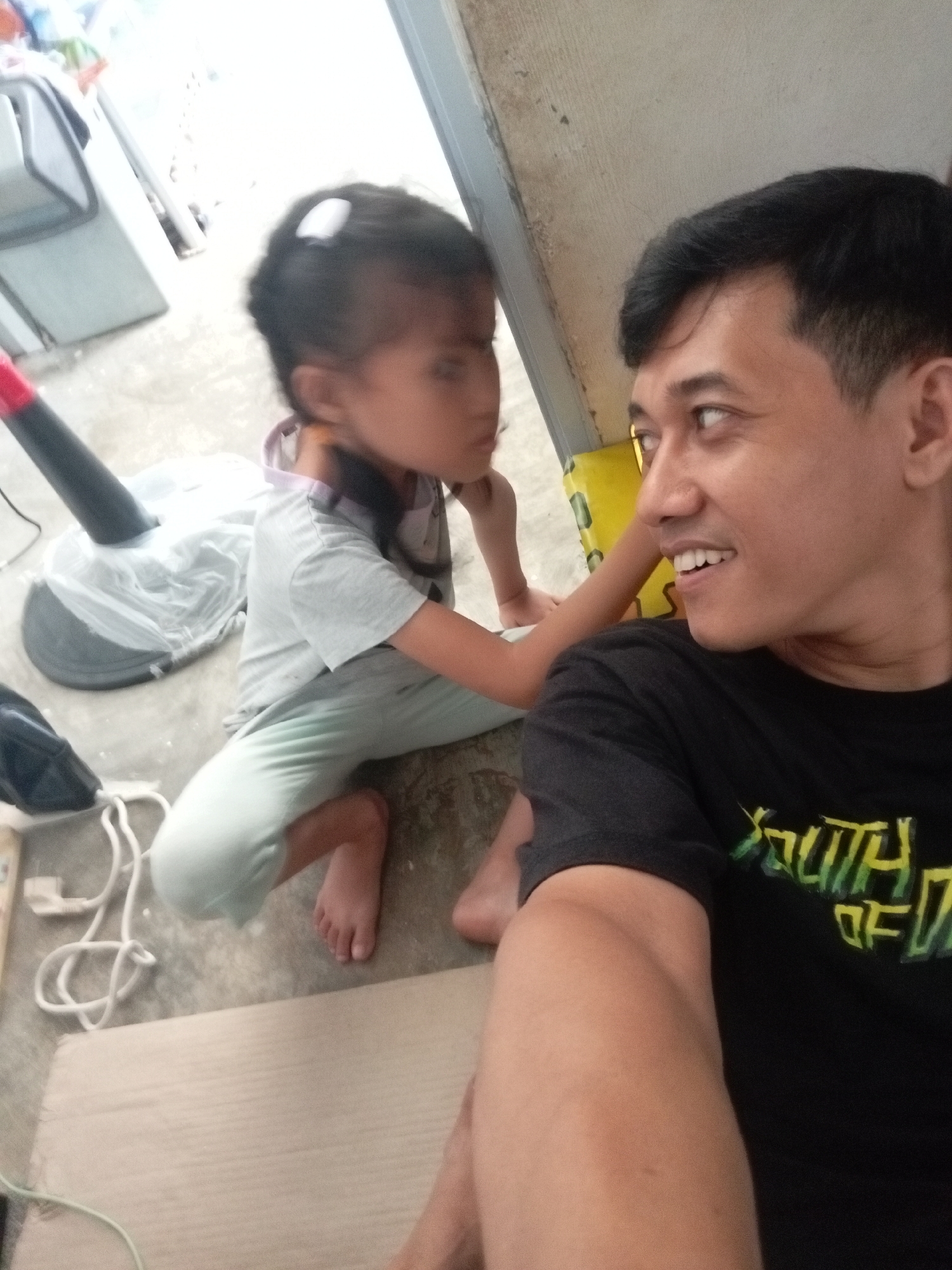 A man and a child are on the floor, looking at each other. The man is wearing a black t-shirt with a yellow and green design on the front. The child is wearing a light gray t-shirt and light blue pants, and has a white clip in her hair. In the background, there is a black floor fan covered in plastic, and some white electrical cords. The scene appears to be indoors, possibly in a home in Batam, Indonesia, given the context.