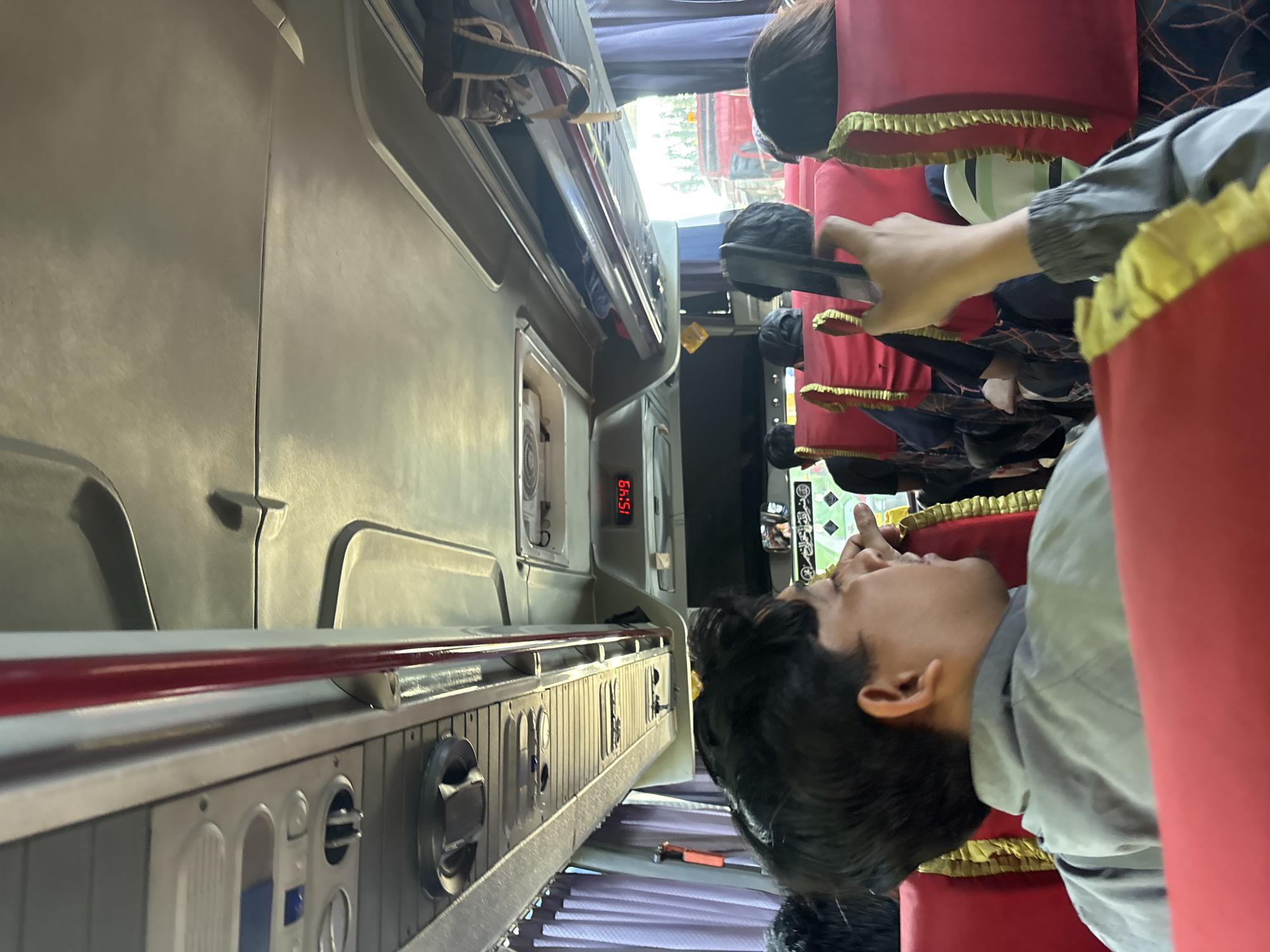 The image shows the interior of a bus. In the foreground, a man is seen from the back, wearing a gray jacket, and holding a phone to his ear. He is seated on a red bus seat with gold trim. Behind him, other passengers are visible, also seated on red seats. The bus ceiling has overhead compartments and a digital clock displaying "15:49". The curtains in the background are purple. The scene appears to be a daytime journey. The presence of the clock and interior of the bus suggests this image was taken within the city of Purwakarta, Indonesia, during transit.