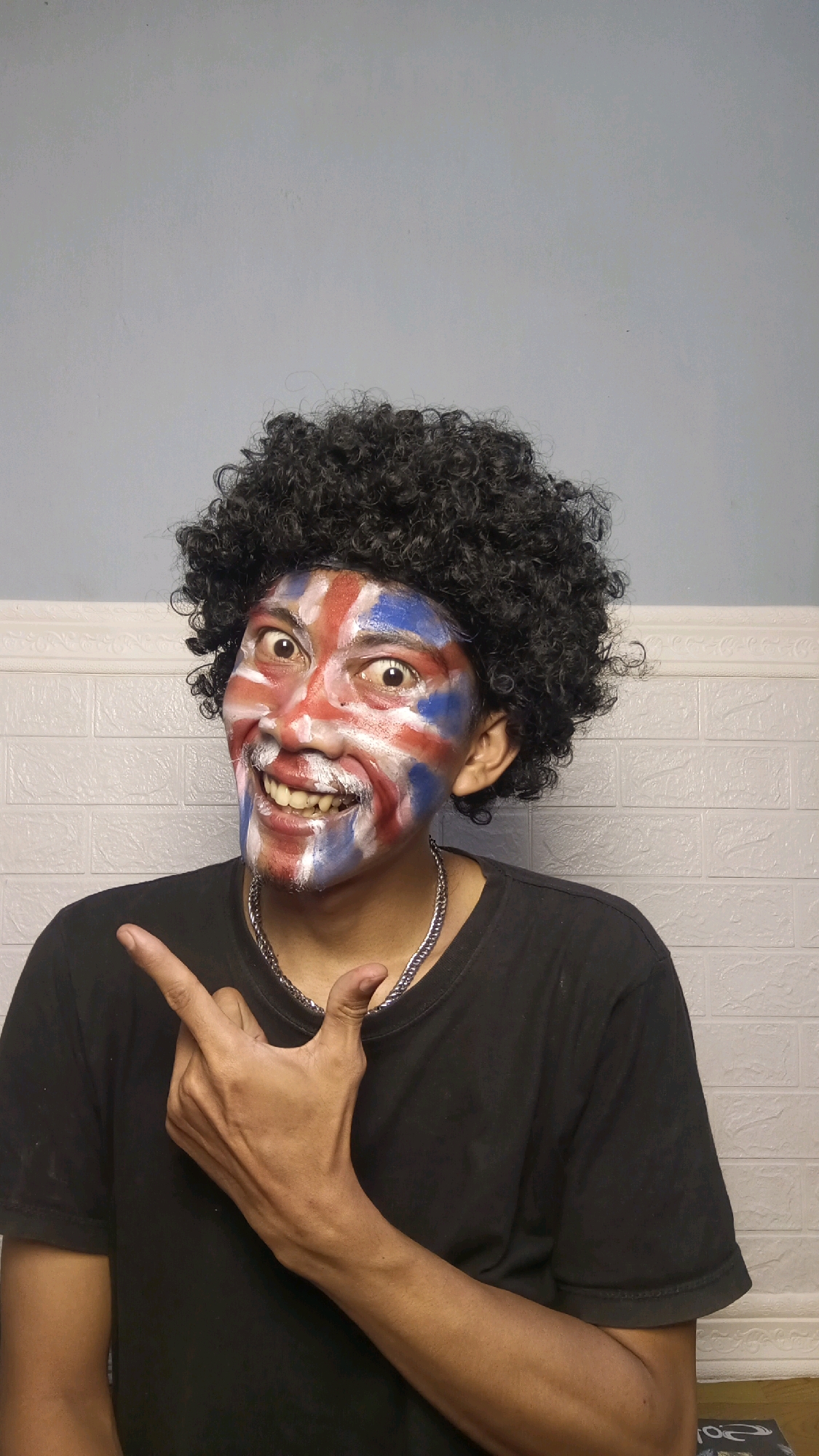 A person with a black afro wig and face paint resembling the Union Jack flag, which is the national flag of the United Kingdom, poses for a photo in a room with a white brick-patterned wall. The person is wearing a black t-shirt and a silver chain necklace. Their left hand is raised, with the index finger pointing to the upper left. The background appears to be indoors, possibly a residence, with a light blue wall visible at the top. There is no indication of the location being in Kebumen, Indonesia.