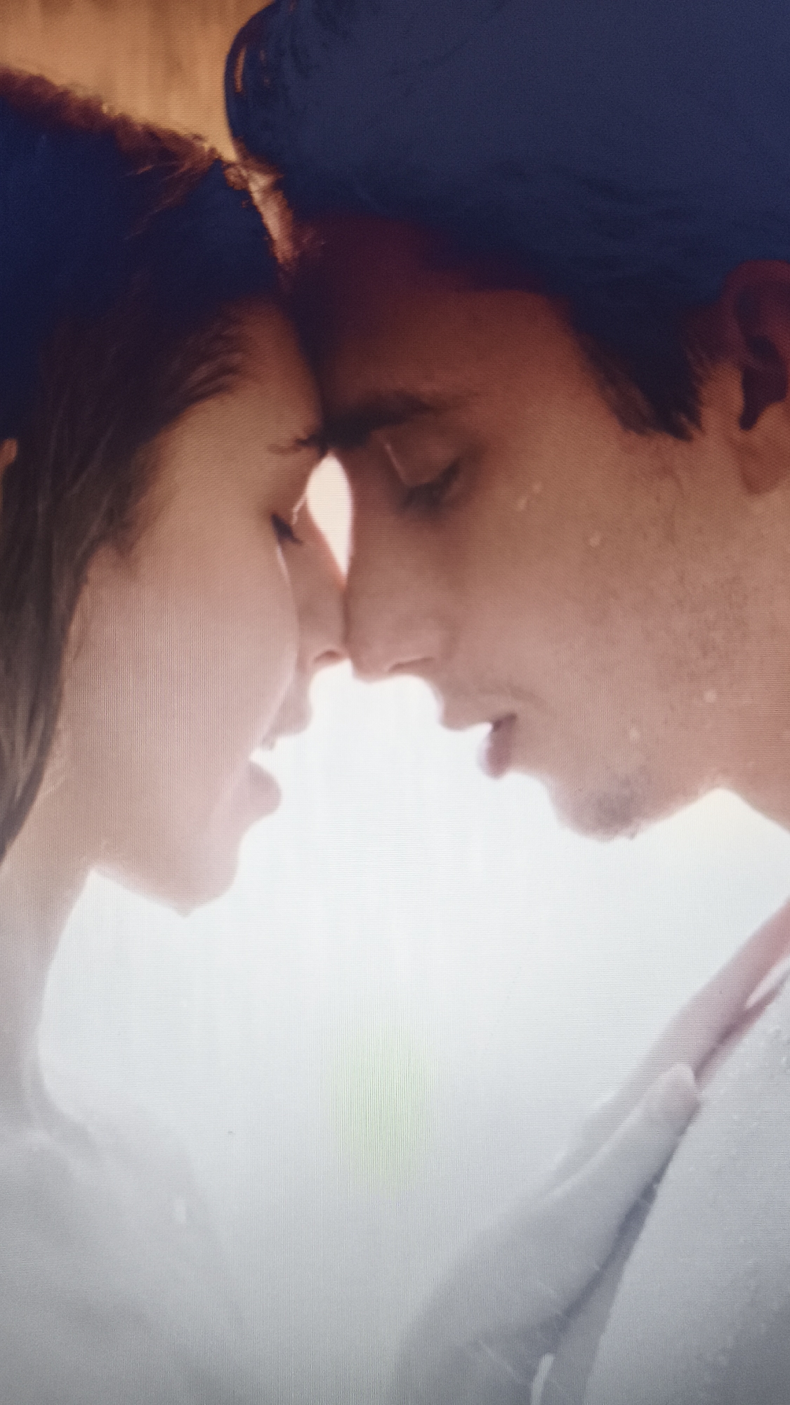 A close-up image depicts a man and a woman facing each other, their foreheads touching. Both have wet hair and appear to be wet, possibly from being in water or rain. The man is on the right, his eyes closed, and appears to have stubble on his face. The woman's eyes are also closed. A hand is visible on the man's chest. The background is bright and somewhat obscured. This image is unlikely to be in Nowy Tomyśl, Poland, as it does not feature any identifiable landmarks or contextual elements specific to that location.