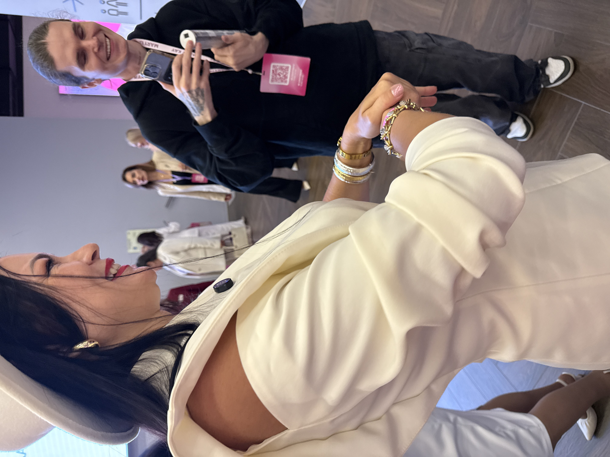 In Moscow, Russia, at an indoor event, a woman with long dark hair, wearing a white hat and an off-the-shoulder white jacket, smiles and looks to her right. She is adorned with gold earrings and multiple bracelets on her left wrist. Next to her, a man with light brown hair pulled back and tattoos on his arms is holding a smartphone and a cylindrical object in his hands, looking towards the woman and smiling. He is wearing a black hoodie and dark pants. In the background, several other individuals are visible, some in business attire, and a pink digital display is mounted on the wall. The flooring appears to be a wooden tile.