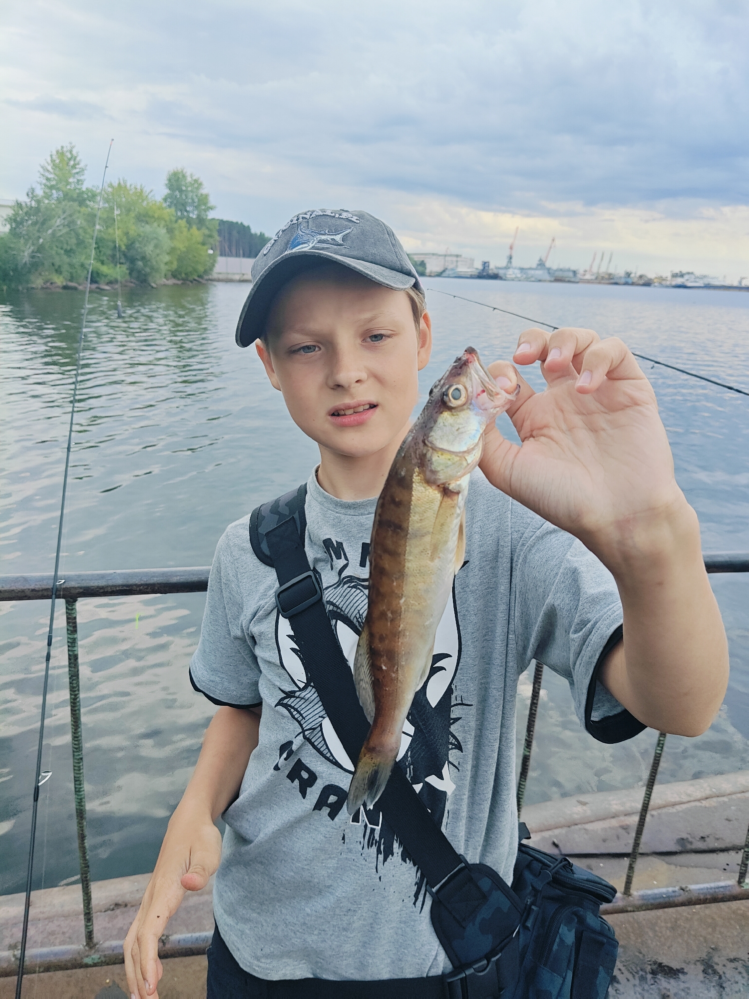A young boy wearing a grey t-shirt and a baseball cap is holding a small fish he has just caught. He is standing on a pier with fishing rods in the background. The water appears to be a river or a bay, with industrial structures visible on the opposite shore. The sky is overcast with clouds. The boy is looking at the camera with a slight smile. The image seems to be taken in Chaikovsky, Russia, as suggested by the industrial landscape often associated with the Kama River in that area.