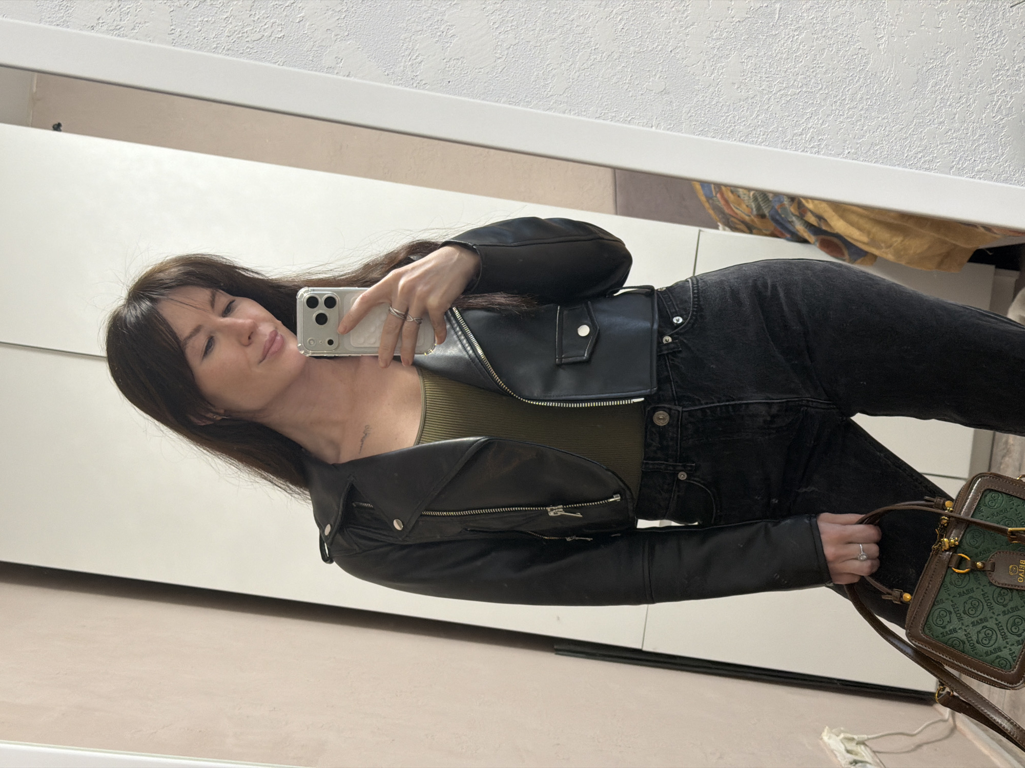 A woman stands indoors in front of a large mirror, taking a selfie. She is wearing a black leather jacket over a green top, dark jeans, and holding a patterned green and brown purse. She has dark hair with bangs. The background is a minimalist interior with white walls and furniture. There is no indication of the city of Perm, Russia in the image.