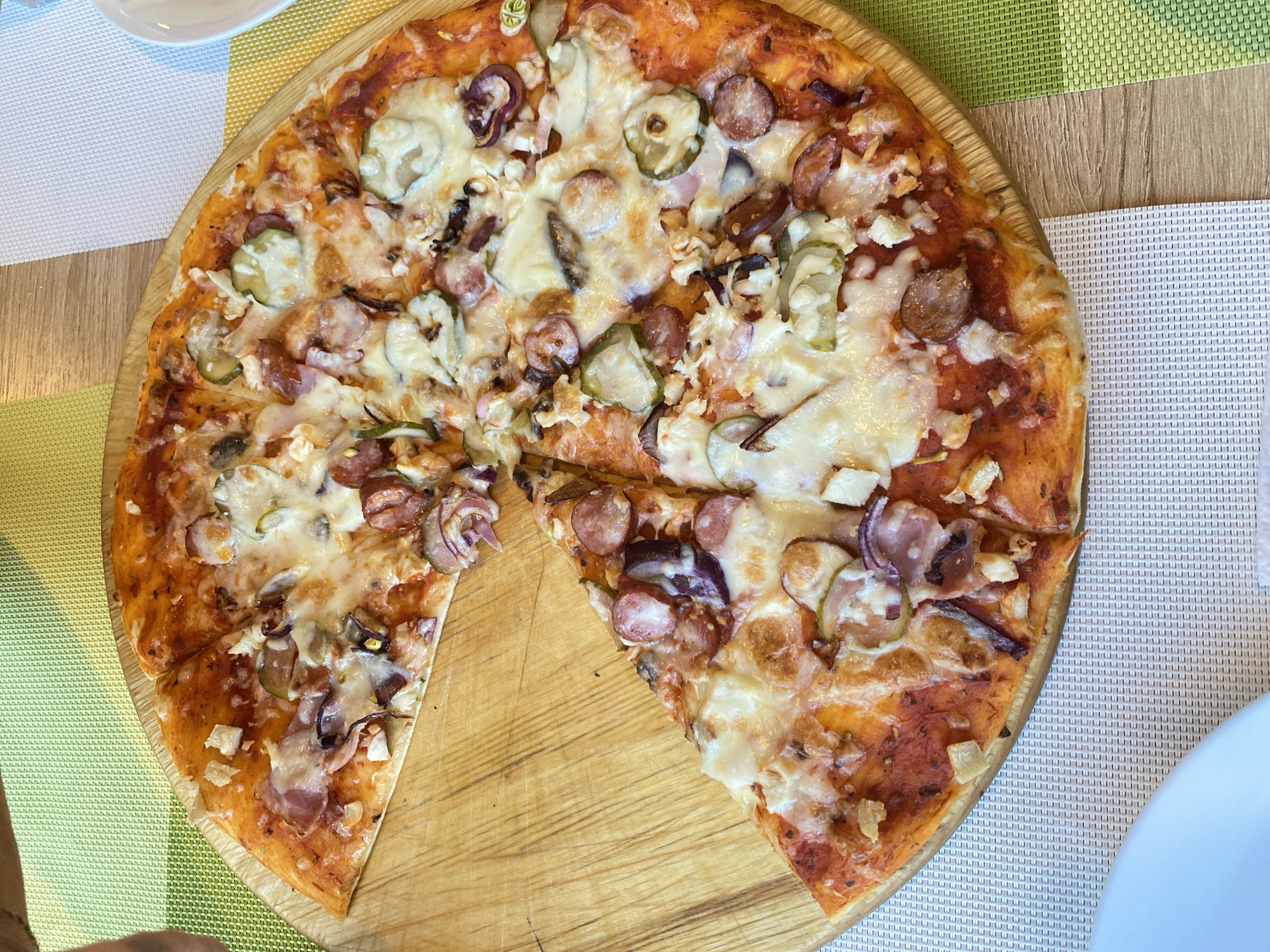 The image displays a partially consumed pizza served on a round, light-colored wooden board. The pizza, which is missing one large slice from its left side, features a red tomato sauce base, melted white cheese, slices of red onion, small round sausage slices, chunks of bacon or ham, and circular slices of pickled cucumbers (gherkins). The remaining pizza is cut into individual slices, forming a nearly complete circle. The wooden board rests on a table covered by two interwoven-textured placemats: a light green one on the left and upper right, and a white/light grey one on the lower right. A portion of a white plate is visible in the lower-left corner of the frame. A faint shadow, possibly from a person's arm or hand, is cast in the upper-left area. The specific location within the city of Chaikovsky, Russia, cannot be determined from the visual information provided.