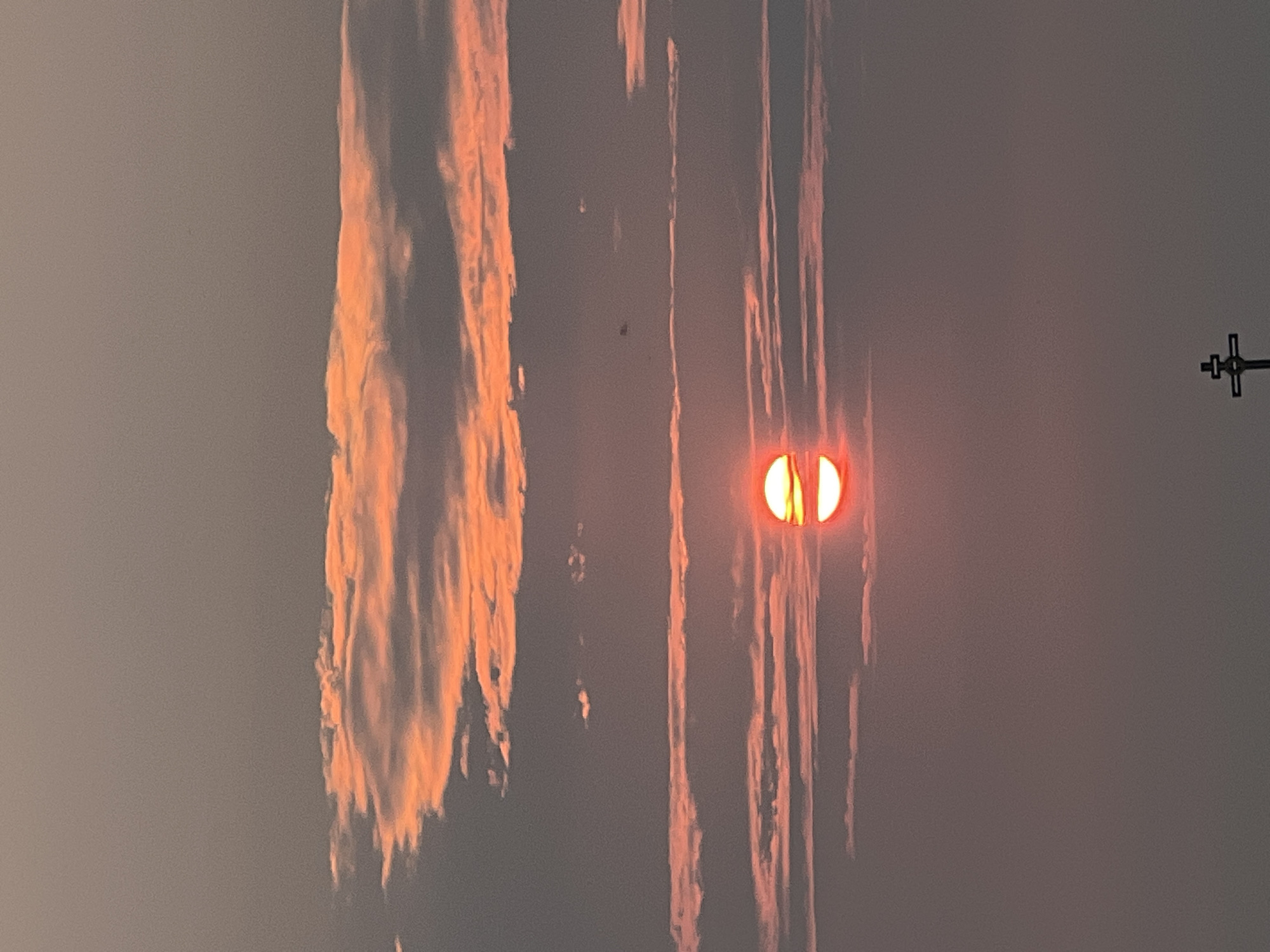 The image depicts a sunset over Novopichugovo, Russia. The sun, a bright orange orb, is partially obscured by horizontal clouds that are illuminated with vibrant orange and pink hues. Above and below the sun, more clouds stretch across the sky, some appearing as thin, wispy lines, while others are more dense. The sky is a gradient of muted grays and purples, transitioning towards a lighter tone at the top of the frame. In the lower portion of the image, a silhouette of a cross is visible against the dusky sky, suggesting a religious or memorial site. The overall atmosphere is serene and tranquil, characteristic of a sunset.