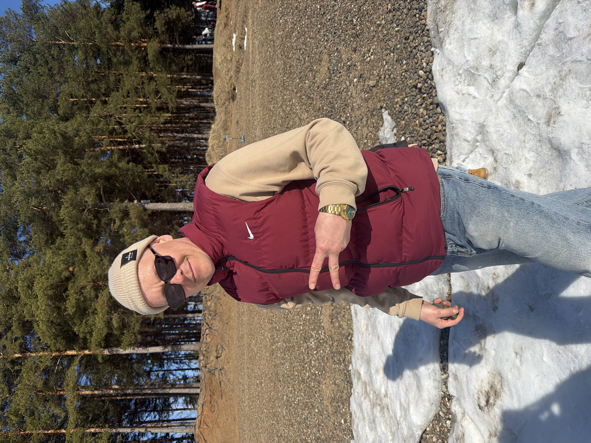A man stands outdoors in a wooded area with patches of snow and gravel ground cover. He wears a beige beanie, sunglasses, a beige sweater, a maroon Nike vest, and blue jeans. A gold watch is visible on his left wrist. He is making a "peace" sign with his left hand and holding a small rock in his right. A dense group of pine trees stands to his left, while a partially visible snowy area lies to his right. The location is in the city of Chaikovsky, Russia.