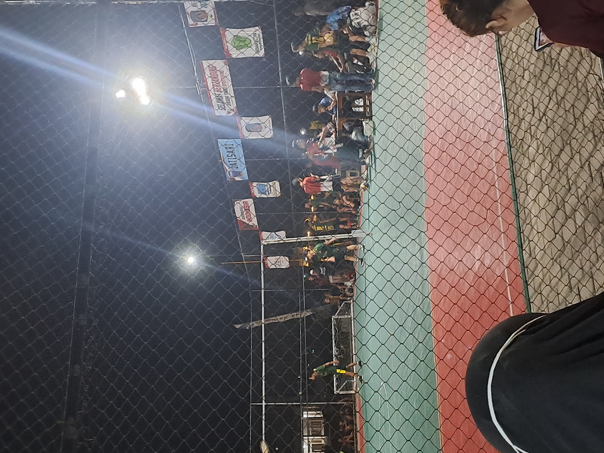 The image depicts a night-time scene at an outdoor futsal court in Sumberejo, Indonesia. A group of young men are playing futsal under bright stadium lights, visible through a chain-link fence. Several spectators are seated or standing behind the fence, watching the game. Banners and flags are strung across the top of the fence, some with text in Indonesian. The court has a green artificial turf surface with red boundaries. In the foreground, a person is partially visible, appearing to be seated and looking at a phone, also behind the fence. The overall atmosphere suggests a casual sporting event or competition.
