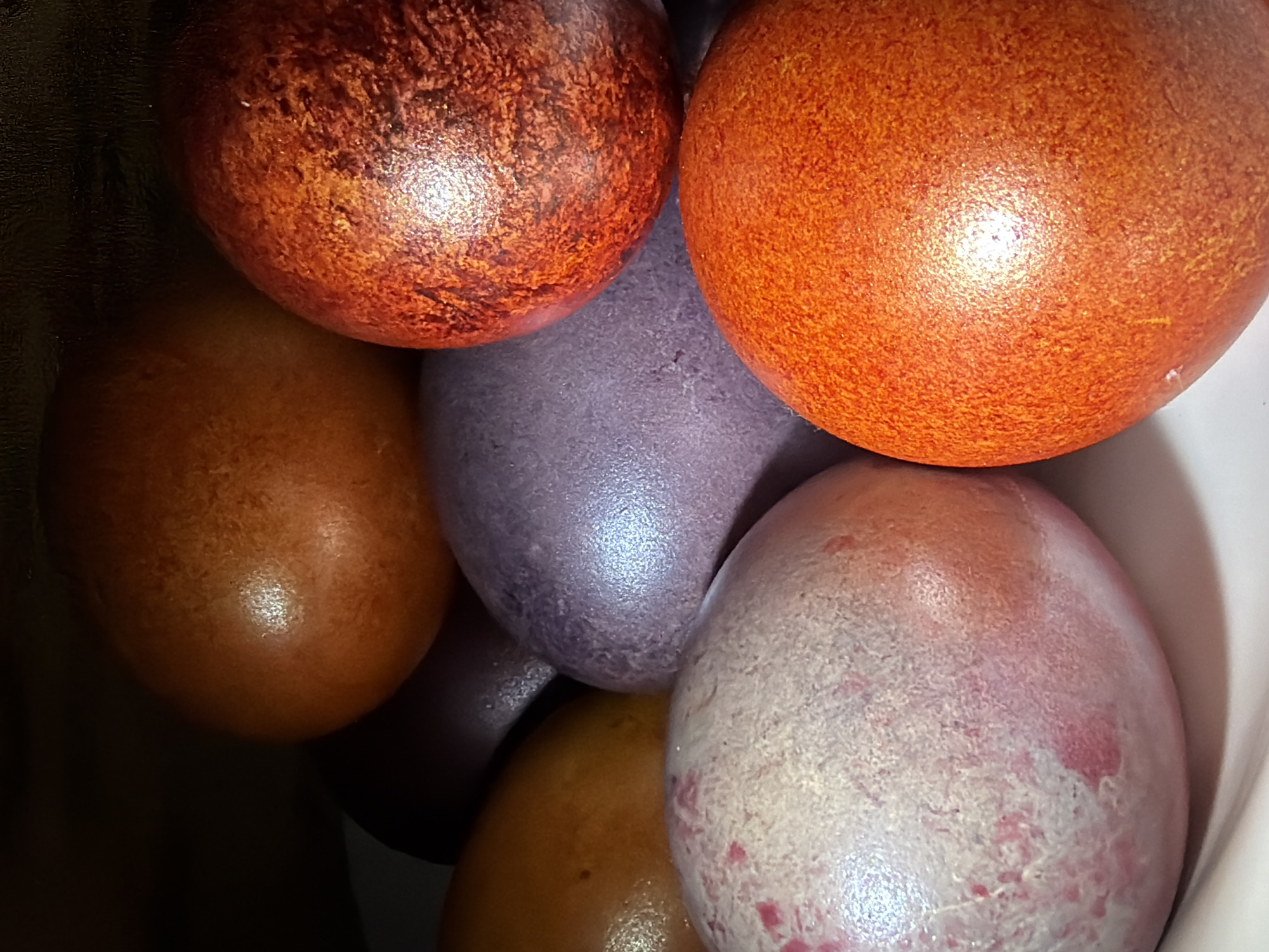A high-angle, close-up shot shows a cluster of six colorful, shiny Easter eggs nestled together in a bowl. The eggs have a variety of colors and surface textures. One egg is a mottled reddish-brown; one is a uniform orange; one is a shiny, smooth brown; one is a smooth purple, and one is a mottled white and pink. The bowl has a white interior.
There is no evidence suggesting that the picture was taken in the city of Chaikovsky, Russia.