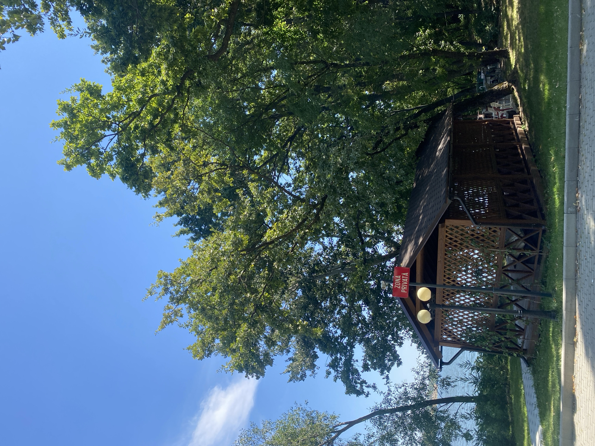 In the city of Ustia, Moldova, a wooden gazebo with a lattice design stands on a grassy area next to a paved walkway. The gazebo has a dark-colored shingled roof and is partially covered in ivy. A sign with red lettering that reads "ZONA PRIVATA" is mounted on a lamppost in front of the gazebo. Two yellow lamps are attached to the lamppost. Beyond the gazebo, a body of water, likely a lake or river, is visible, bordered by trees and a shoreline. Lush green trees with dense foliage dominate the background, with a clear blue sky above. The scene appears to be a tranquil outdoor setting.