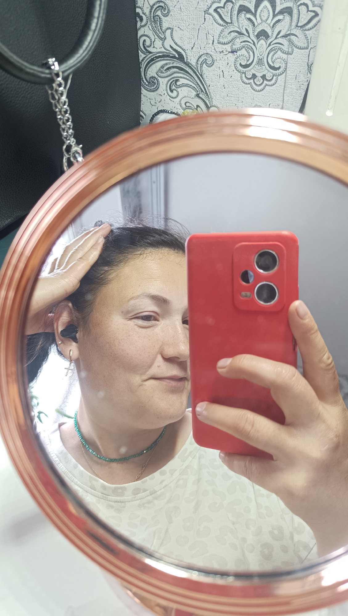 In an indoor setting, a woman is taking a mirror selfie. She is positioned in front of a mirror with a rose gold colored frame, which reflects her image. She is wearing a white t-shirt with a leopard print pattern, a green beaded necklace, and a gold chain. She has black wireless earbuds in her ears and a cross earring in her left ear. Her right hand is raised, holding a red smartphone in front of her to capture the reflection. The background of the reflection shows a wall with a floral patterned wallpaper. To the left of the mirror, a black leather bag with a silver chain is visible. The scene appears to be in a residential environment within the city of Chaikovsky, Russia.