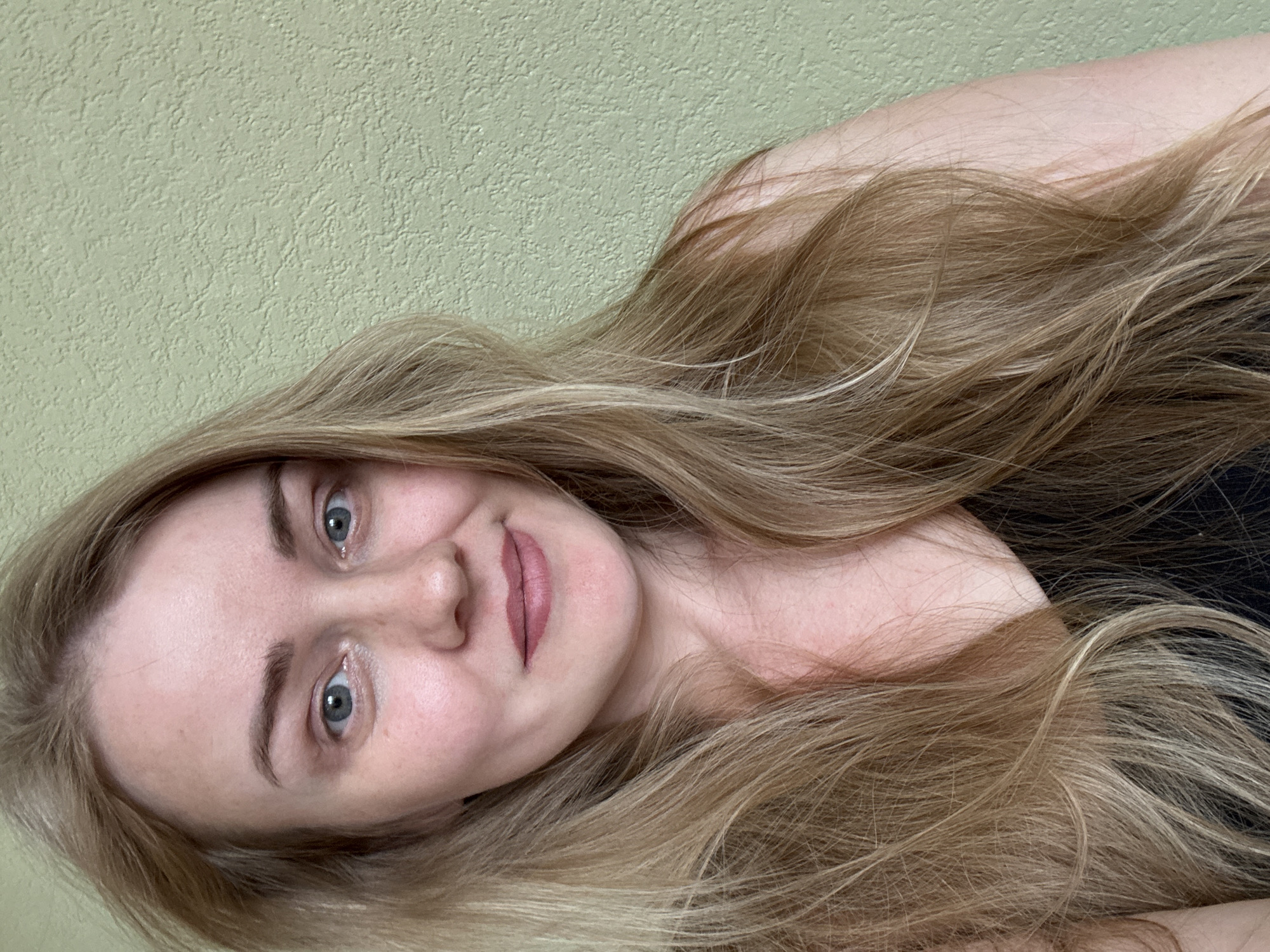 A portrait of a fair-skinned woman with blonde hair and blue eyes is captured against a light green textured wall. The woman is facing the camera with a gentle smile. Her hair is flowing around her face and shoulder. She is wearing a black top. The image does not provide any information to place the woman in the city of Chaikovsky, Russia or any location in the city.
