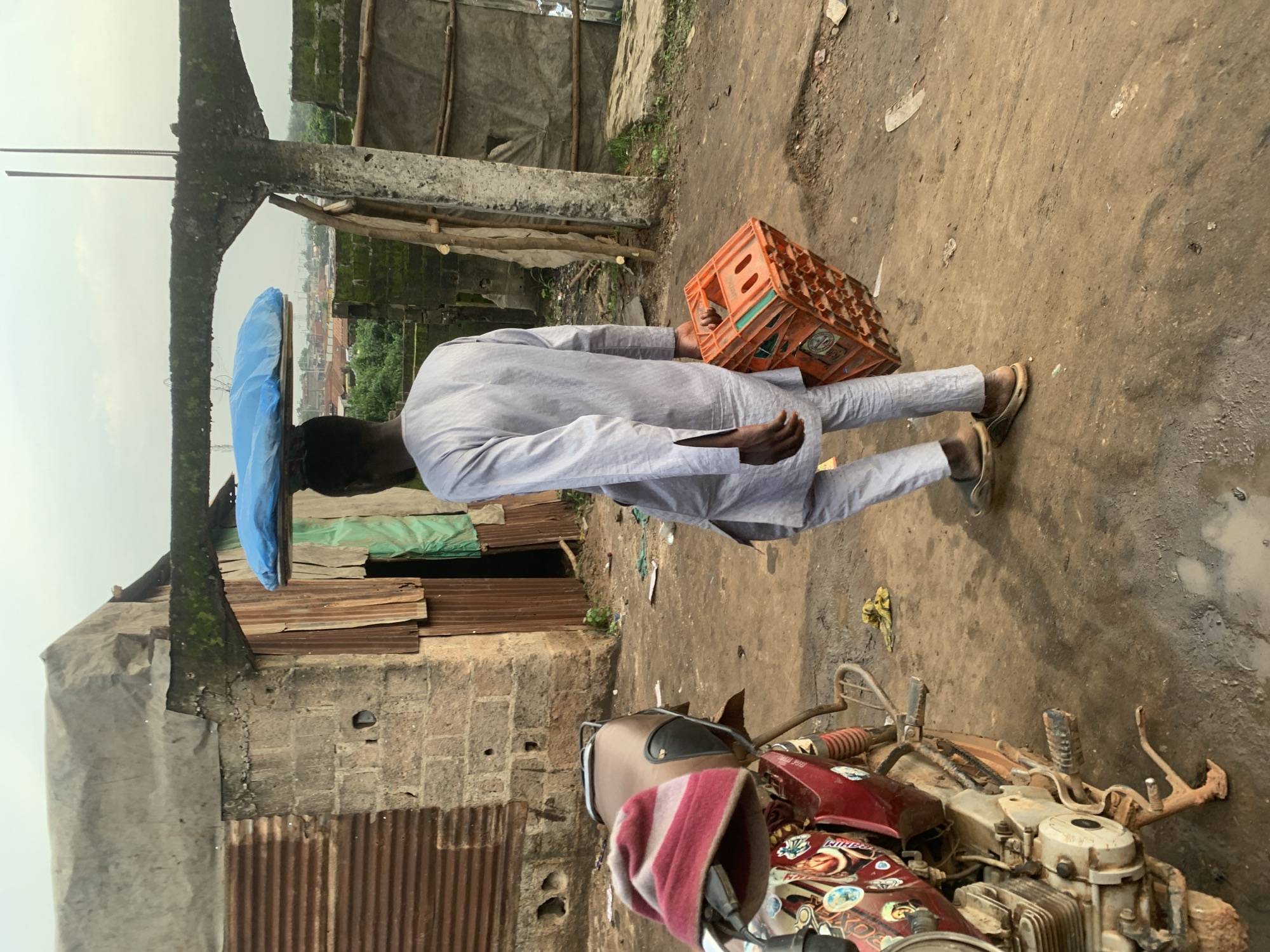 In Shagamu, Nigeria, a Black man walks away from the camera, carrying an orange plastic crate in his right hand and balancing a blue tarp-covered object on his head. He is wearing a long-sleeved, light-colored tunic and matching pants, with open-toed sandals. The man is walking on a dirt path littered with debris, next to a partially constructed building with corrugated metal siding and brickwork. A red motorcycle with a maroon seat and a striped hat on its handlebars is parked to the left of the path. The background shows a hazy cityscape with trees and buildings.