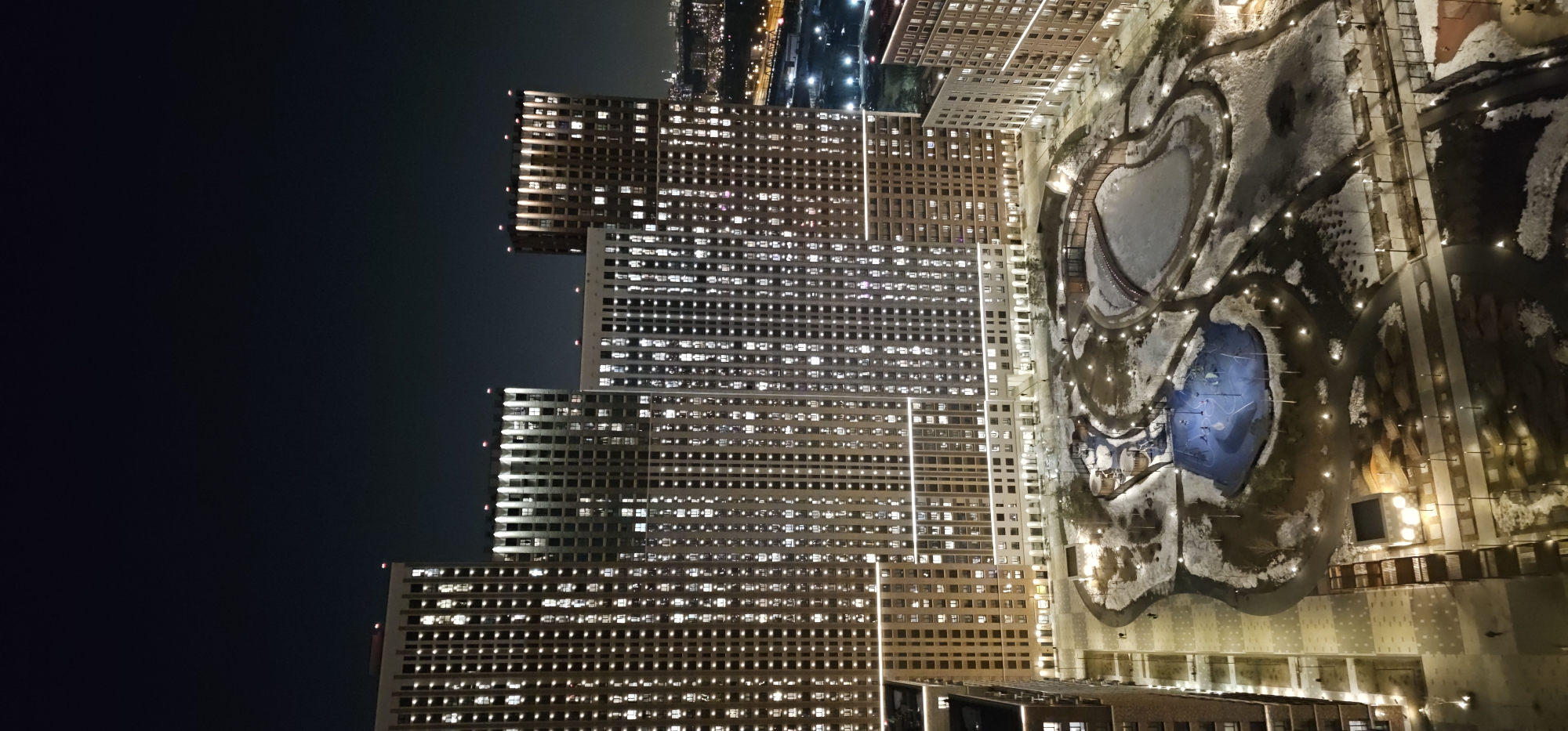 An overhead nighttime image depicts a modern urban residential complex, identified as part of the ZilArt development in Moscow, Russia. The scene is illuminated by extensive artificial lighting.
Dominating the background are multiple high-rise residential buildings, exhibiting a contemporary architectural style with light-colored facades and numerous windows. A significant number of windows are illuminated, indicating occupancy. Some building rooftops feature red lights, possibly aviation obstruction markers.
The foreground and midground feature a landscaped park or courtyard area situated between the buildings. Patches of snow and bare ground are visible, suggesting winter or early spring conditions. Winding pathways traverse the park, clearly delineated by a dense array of uniformly spaced, low-level streetlights.
A central feature of the park is a large, irregularly shaped body of water, currently frozen over. A dark-toned wooden pedestrian bridge arches across one section of this frozen area. Adjacent to the frozen pond is a distinct, circular play area, brightly illuminated with a blue light, featuring various play structures. Scattered bare trees and shrubs populate the park.
A few human figures, appearing as small silhouettes, are discernible walking on the illuminated pathways. In the far distance, to the right, a faint urban skyline with scattered lights and a distinct illuminated bridge structure are visible against the dark night sky. The overall impression is one of a well-maintained and active residential area at night.