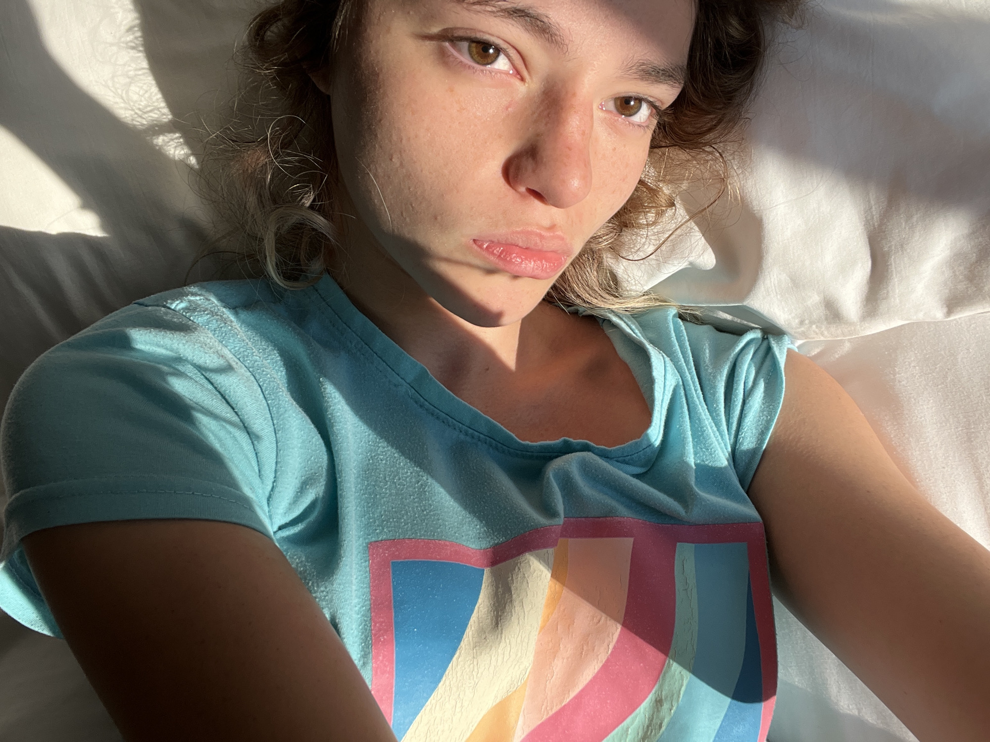 A young woman with light skin and brown eyes, lying on a white pillow, is taking a selfie. She has brown hair with blonde highlights and freckles on her face. She is wearing a light blue t-shirt with a colorful graphic design. Light is illuminating her face and creating shadows on her shirt and the pillow. The location cannot be determined from the image.