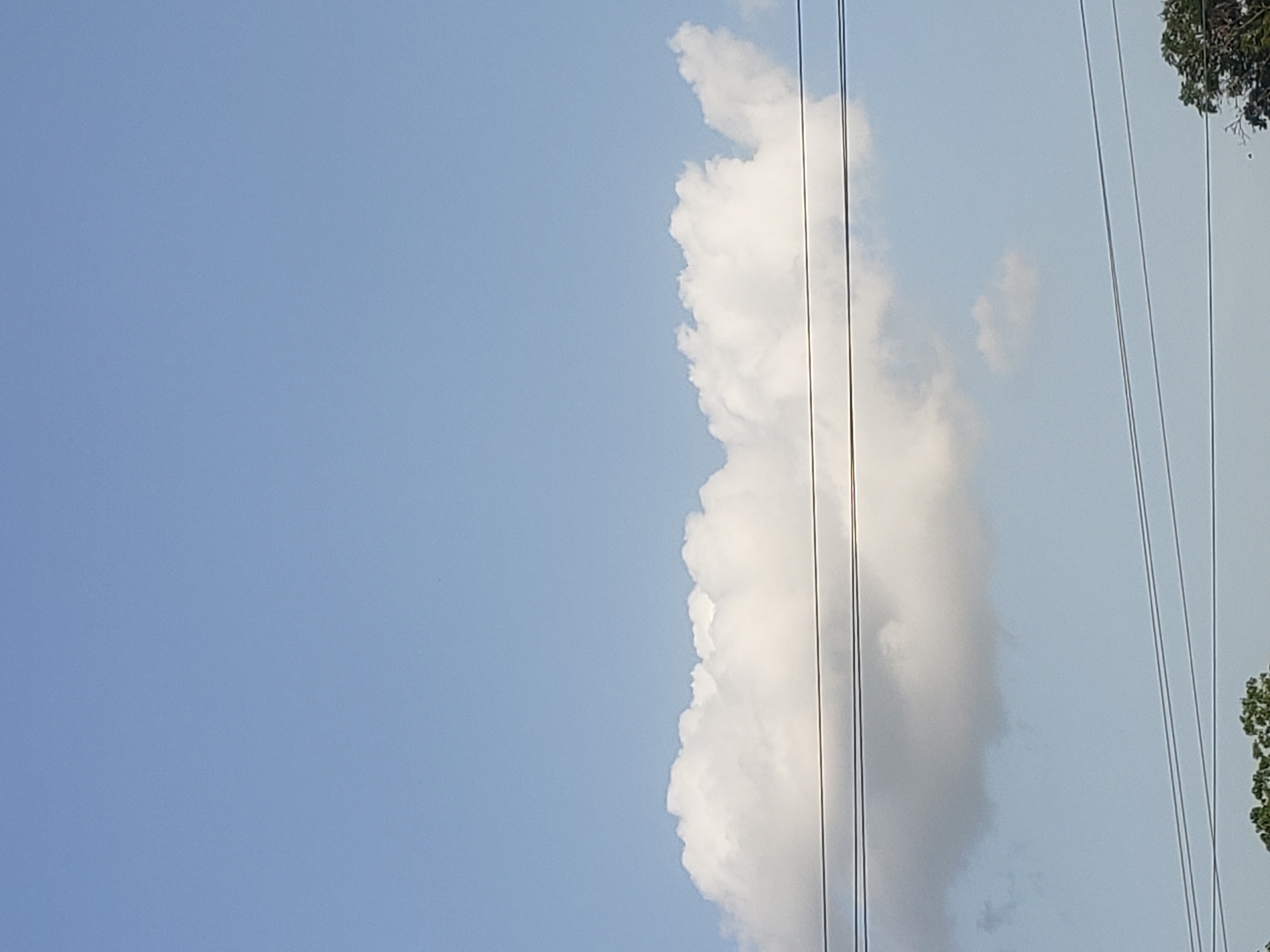 The image shows a blue sky with a large, white cumulus cloud on the right side of the frame. There are several power lines running vertically through the cloud. The top right corner of the image has tree branches and foliage. The image was taken in the city of Adankolo, Nigeria.