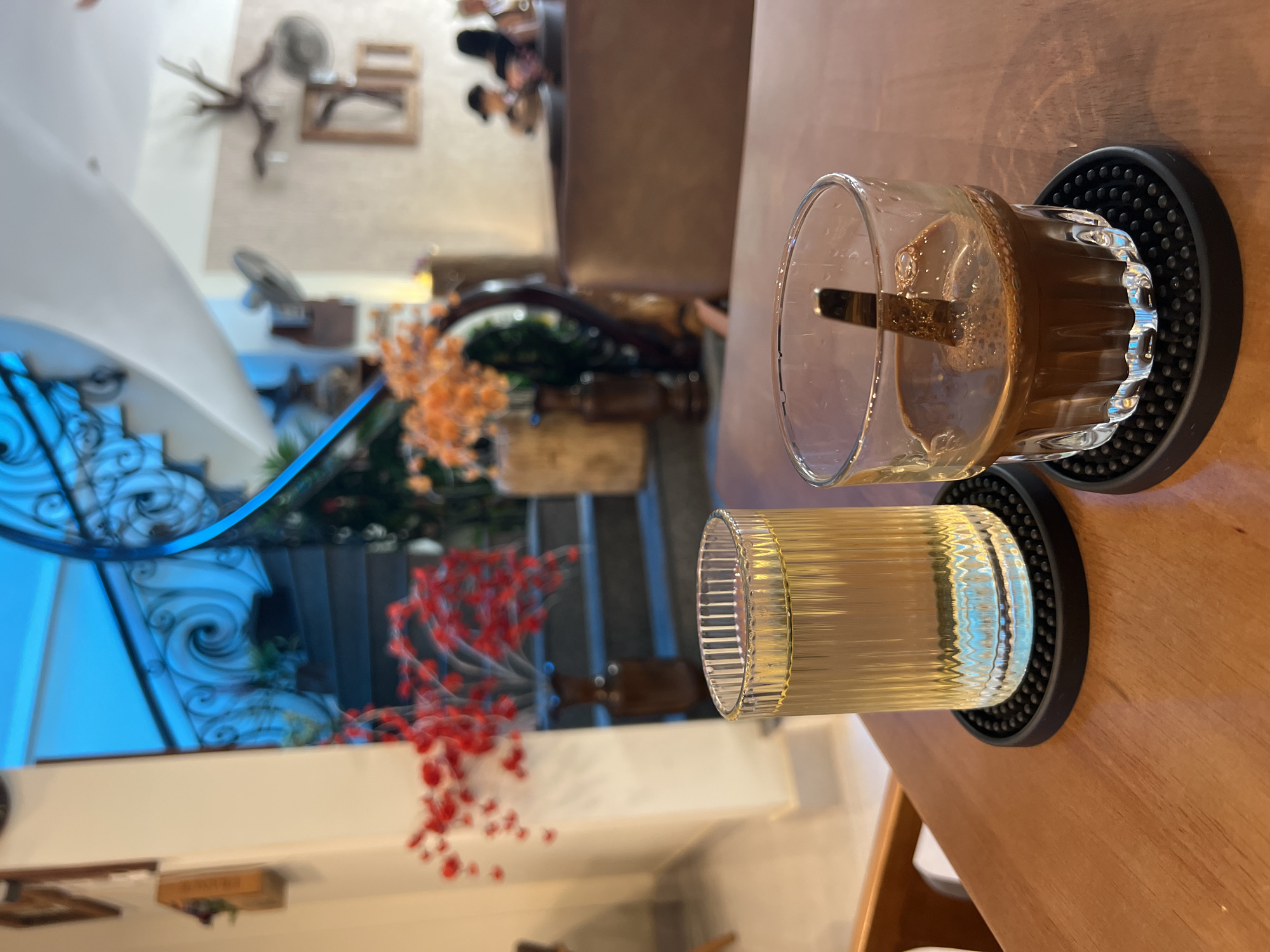 The image depicts a close-up of two glasses on a wooden table. One glass contains a dark brown liquid, possibly coffee, with a spoon visible inside. The other glass is filled with a clear, yellowish liquid, possibly water or tea. Both glasses are placed on black, round coasters with a textured surface.
In the blurred background, there is a multi-level interior space with a decorative staircase featuring blue metal railings. Orange and red floral decorations are visible near the staircase. Further back, there are hints of furniture and other decor, suggesting a cafe or restaurant setting.
The image does not provide enough information to determine the exact location or any details about people, actions, or interactions.