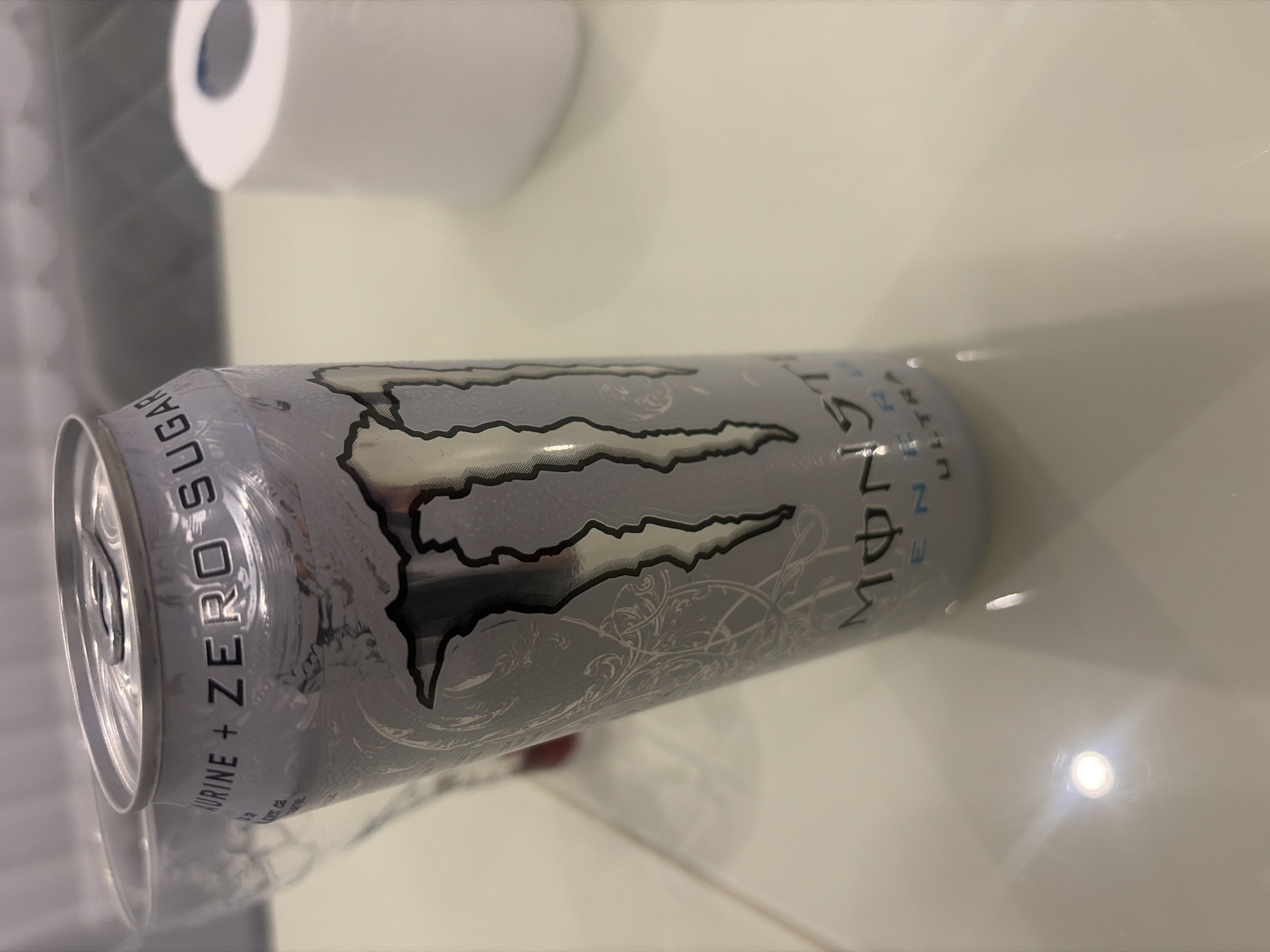 A Monster Energy Ultra can is the central subject, positioned at an angle on a light-colored, reflective surface. The can is silver with the iconic Monster logo in black and silver. Text visible on the can includes "TAURINE + ZERO SUGAR" and "MONSTER ENERGY ULTRA". To the left of the can, a portion of a clear glass is visible. In the background, a white roll of what appears to be toilet paper sits on the same surface. The lighting suggests an indoor setting, possibly a table. The scene is static and appears to be a product shot or still life. There is no indication of a specific location in Podgumer, Bulgaria.