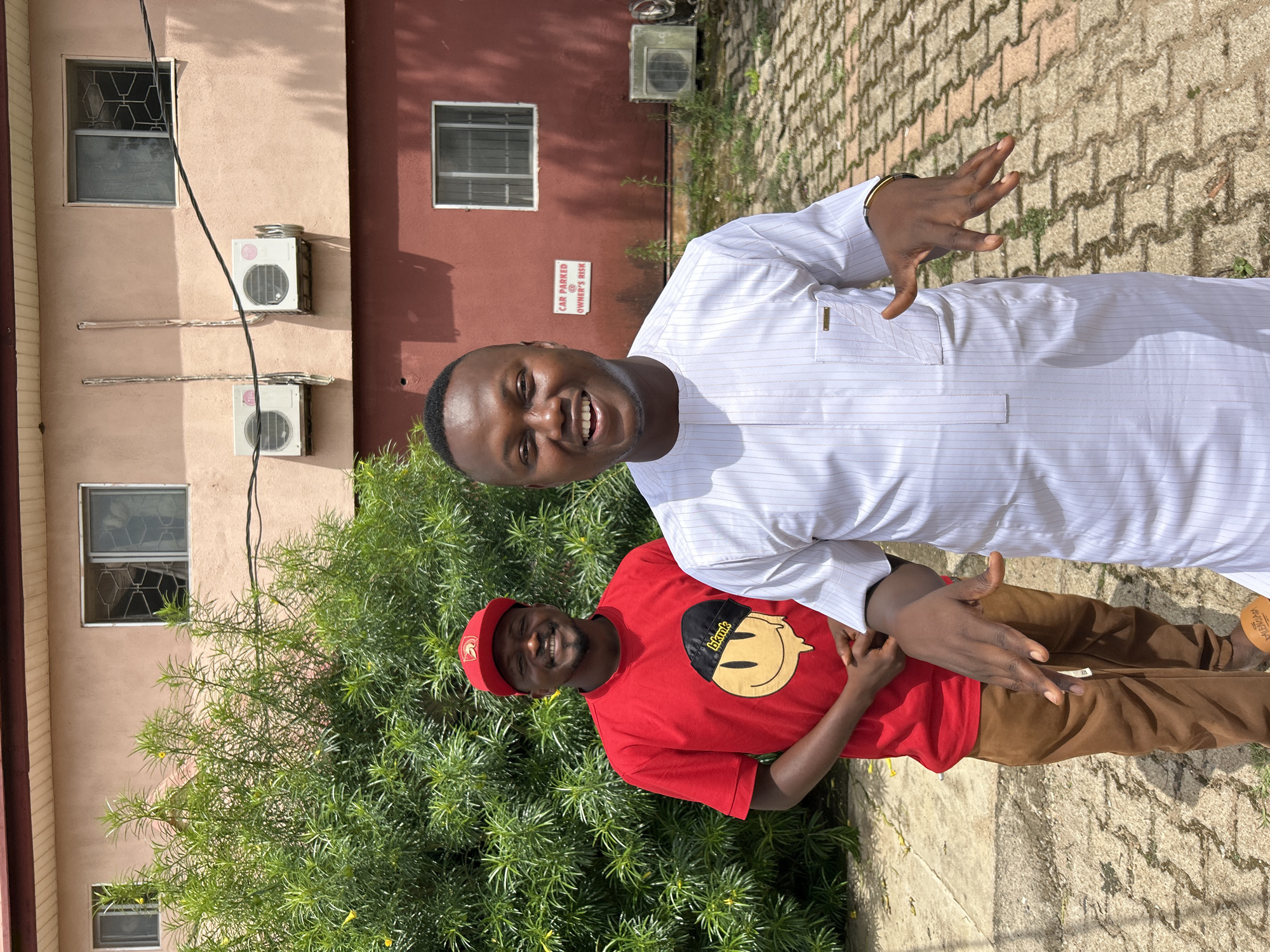 The image depicts two men standing outdoors in a brightly lit environment, presumably in Jos, Nigeria.
In the foreground, slightly to the right of the frame, a man with dark skin, short dark hair, and a broad smile is visible. He is wearing a white, vertically striped traditional tunic (agbada-like top) with a small gold-colored emblem on the left chest pocket. His hands are slightly raised and open, with fingers splayed, as if gesturing or in mid-sentence. A gold-colored bracelet is visible on his right wrist.
Behind and slightly to the left of the first man, a second man with dark skin is present. He is smiling broadly and wearing a red short-sleeved t-shirt featuring a stylized yellow melting smiley face with a black beanie labeled "bkmk." He also wears a red baseball cap with a gold Spartan helmet emblem on the front. His hands are clasped in front of him. He is wearing brown, loose-fitting trousers and a visible tan sandal on his left foot with the word "MUSTAPHA" printed on it.
The ground beneath them is paved with light brown interlocking concrete pavers, interspersed with small patches of green weeds.
The background features a two-story building with a light peach-colored upper section and a darker reddish-brown lower section. Multiple windows, some with metal security grilles, are visible on both levels. Two white outdoor air conditioning units are mounted on the upper peach wall, with associated piping. A third, older, and discolored AC unit is visible on the lower reddish-brown wall to the far right. A white sign with red text, "CAR PARKED OWNER'S RISK," is affixed to the reddish-brown wall. A thick black electrical cable traverses the upper wall horizontally. To the left and center background, a dense green bush with slender leaves and small yellow flowers partially obscures the building. The sky is bright and clear, indicating daylight.