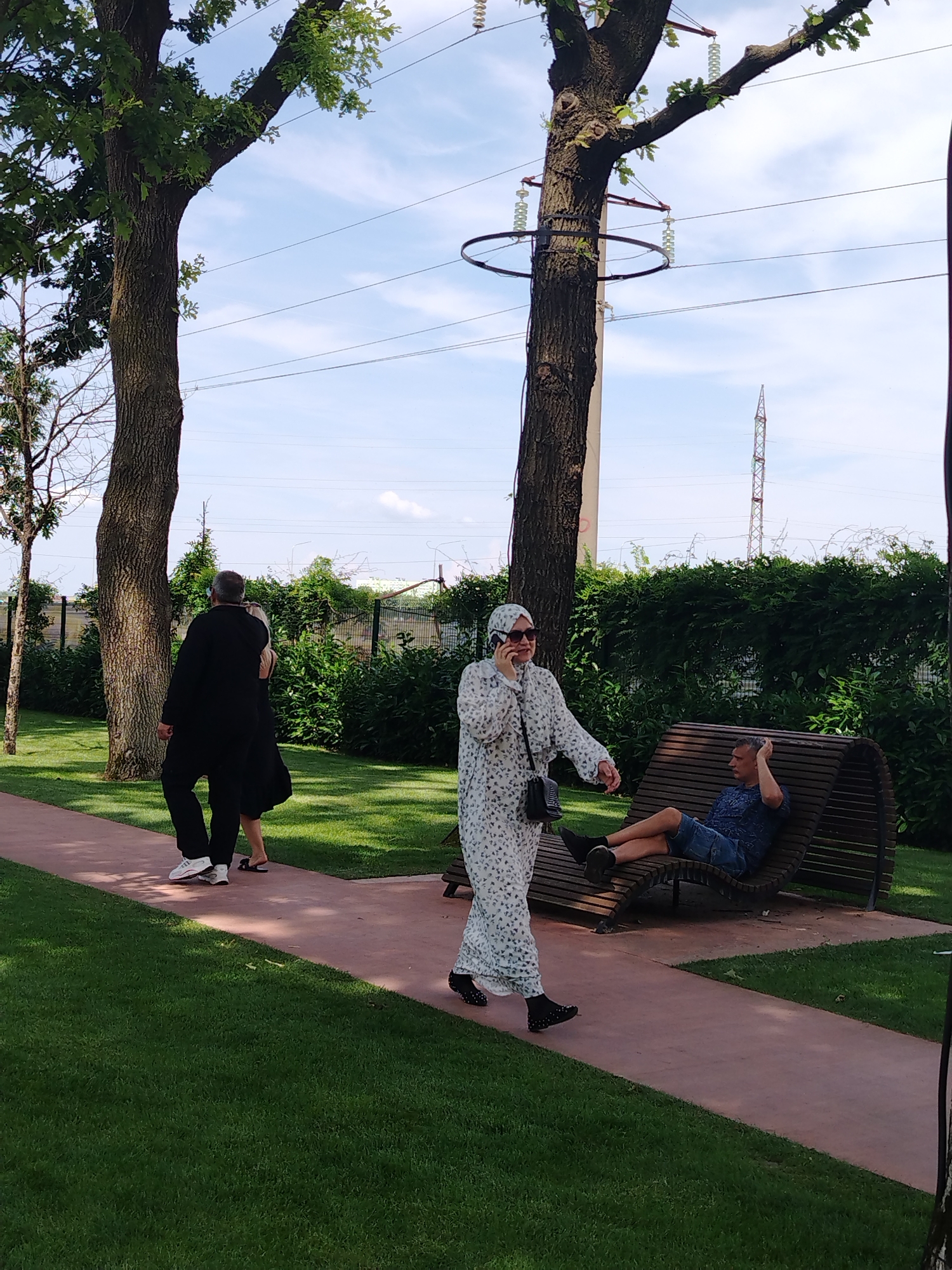 A man in a black suit and white sneakers walks on a pink path in a park with a woman in a black dress. A woman wearing a white patterned dress and a hijab walks by, talking on her phone. A man in a blue shirt and denim shorts relaxes on a wooden bench. The scene is set against a backdrop of green grass, trees, and a clear blue sky with some clouds. In the distance, power lines and a transmission tower are visible. The overall impression is of a public park area in the city of Krasnodar, Russia, on a pleasant day.