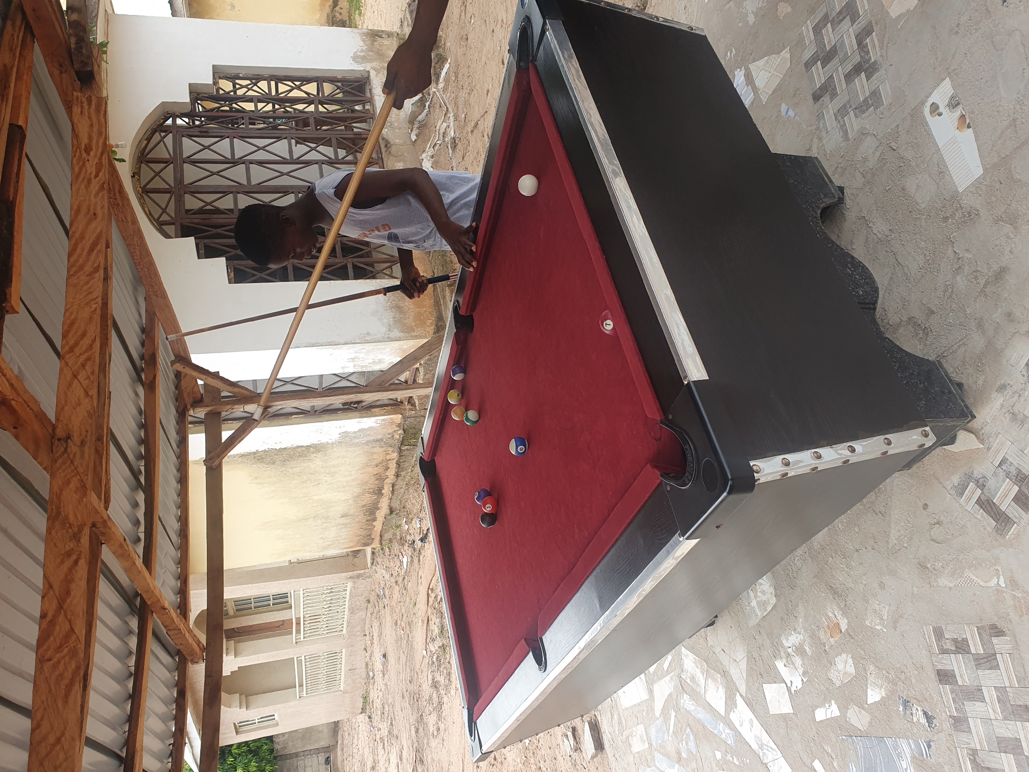 A young Black male in a grey tank top is playing pool outdoors in Lokoja, Nigeria. He is standing behind a red felt pool table with black sides, holding a pool cue. There are several pool balls on the table, including a white cue ball, a striped blue ball with the number 10, and a solid red ball with the number 3. Behind the pool table, a white building with a metal grate on the windows is visible, partially covered by a wooden and corrugated metal roof. The ground is a mix of sand and broken tiles.