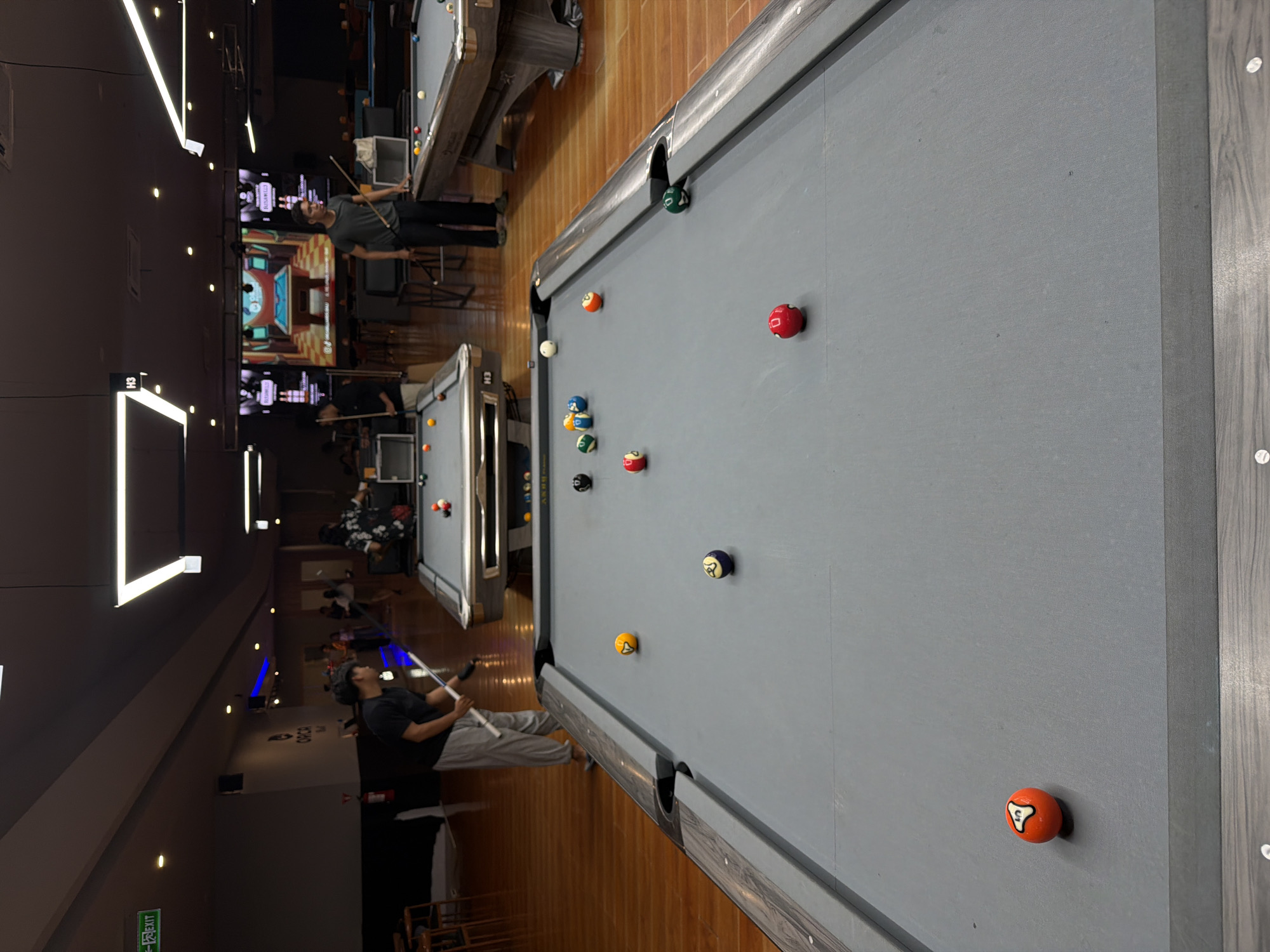 The image captures a pool hall setting in Kuta Selatan, Indonesia. A man in a black shirt and grey pants is positioned on the left side of the frame, holding a pool cue, seemingly preparing to strike an orange ball located on the right side of the pool table. The pool table is gray and has multiple other billiard balls scattered across its surface.
In the background, other pool tables are visible, along with people playing. There are also large screens displaying video game content and bar-style seating. The lighting appears to be a mix of natural and artificial light, with some neon lights present. The flooring is a light brown wood.