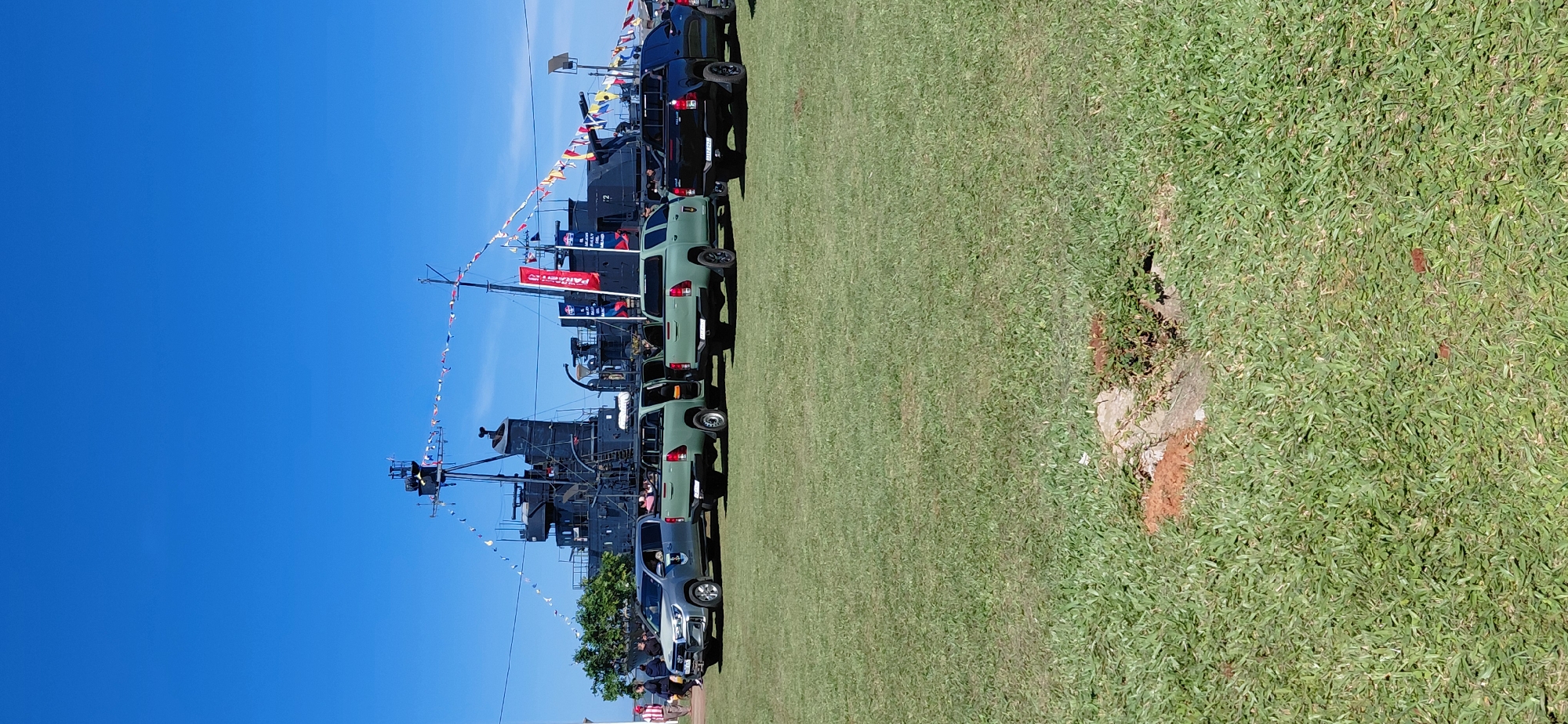 In Encarnación, Paraguay, on a clear day with a bright blue sky, a black naval ship is docked. The ship features a prominent bridge tower and is adorned with a string of colorful flags. In front of the ship, parked on a meticulously manicured green lawn, are four pickup trucks. Three of the trucks are olive green, while one is black. All four trucks appear to be military or official vehicles. To the left of the trucks, a group of people are gathered near the shoreline, with some individuals visible on the ship's deck. The scene suggests an outdoor event or exhibition, possibly a naval open house or a historical reenactment. The foreground is dominated by the green grass, with a small patch of exposed earth and vegetation in the lower center.