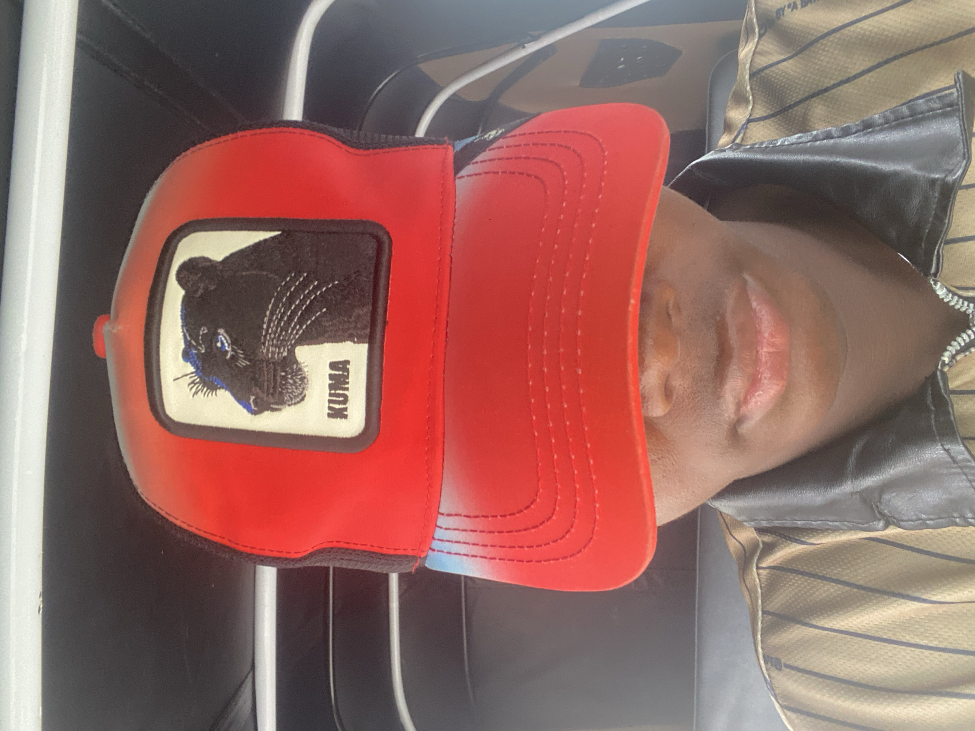 A person wearing a red cap with a black panther emblem and the word "KUMA" is captured in a close-up, head-and-shoulders shot. The cap has a red front panel with a gradient fading to black at the sides and a black mesh back. The black panther image is embroidered on the front. The person's face is partially obscured by the cap's brim, with only their mouth and chin visible. They are wearing a tan and dark striped shirt with a black collar. The background appears to be the interior of a vehicle, possibly a tuk-tuk, with dark upholstery. The lighting suggests an outdoor setting, possibly during the day. The image was taken in the city of Ado, Nigeria.