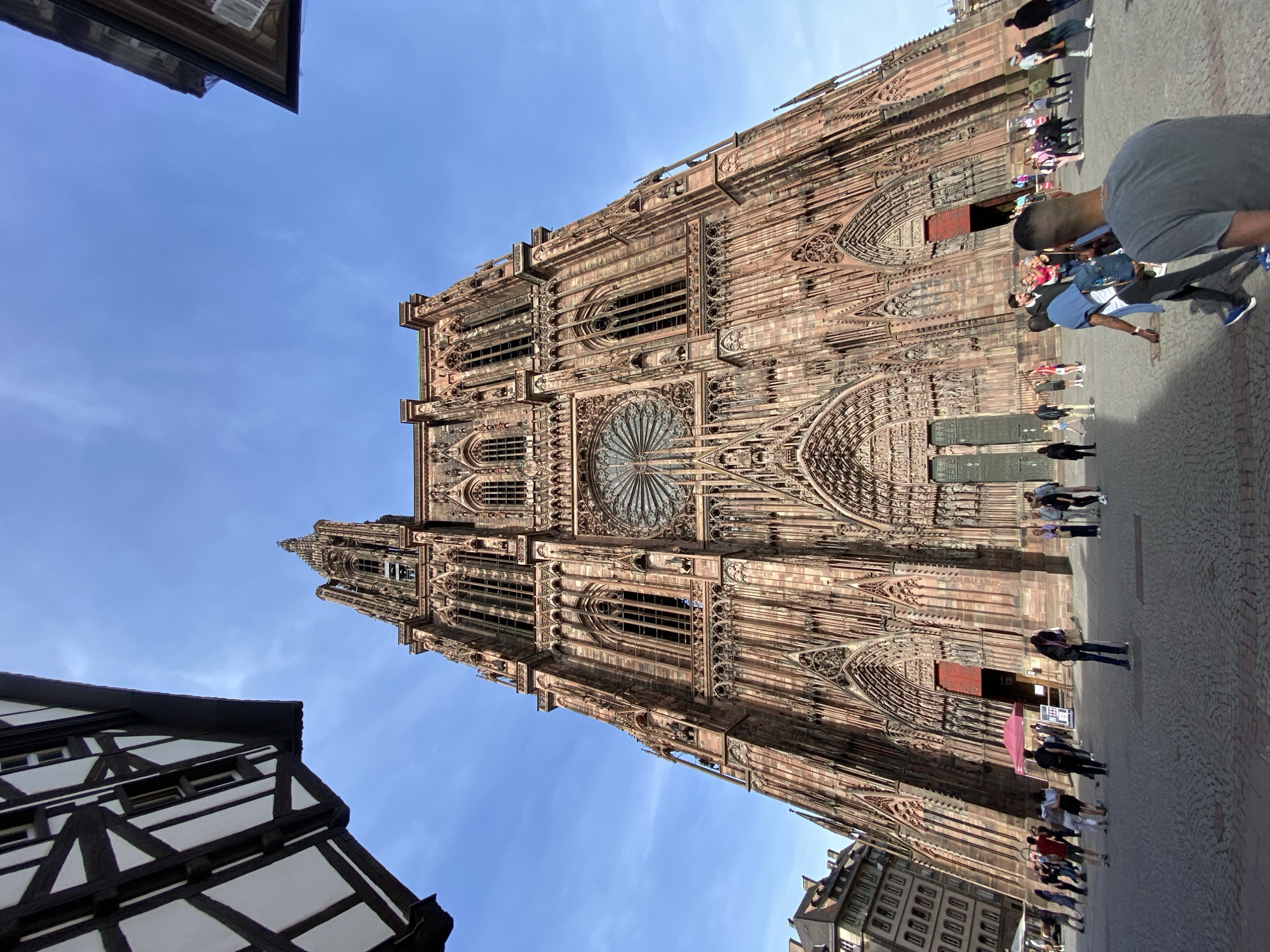 The image, rotated counter-clockwise, presents a majestic view of the Strasbourg Cathedral (Cathédrale Notre-Dame de Strasbourg), dominating the frame from a low angle, looking upwards towards its intricate facade and spires against a clear blue sky.
The cathedral, constructed from reddish-brown sandstone, showcases elaborate Gothic architecture. Its monumental western facade is highly detailed, featuring multiple arched portals with intricate sculptural work in their tympanums and archivolts. A prominent large rose window, characteristic of Gothic cathedrals, is centrally located on the upper part of the facade. Above and around it, numerous lancet windows, pinnacles, and decorative elements add to its grandeur. The tall, slender spires and bell towers rise towards the sky, though the very top of the tallest spire is partially out of frame on the left.
In front of the cathedral is a paved public square, likely cobblestone, where numerous people are gathered. They appear to be tourists or visitors, standing, walking, and observing the cathedral. One person in a grey t-shirt is visible in the mid-right foreground, facing away from the camera. Another traditional half-timbered building with white walls and dark beams, typical of the Alsace region, partially frames the lower left corner of the scene. The image captures a bright, sunny day, suggesting late morning or afternoon, under a clear, vibrant blue sky with no discernible clouds. No clearly legible text is visible within the frame. The scene conveys a busy, tourist-filled atmosphere.