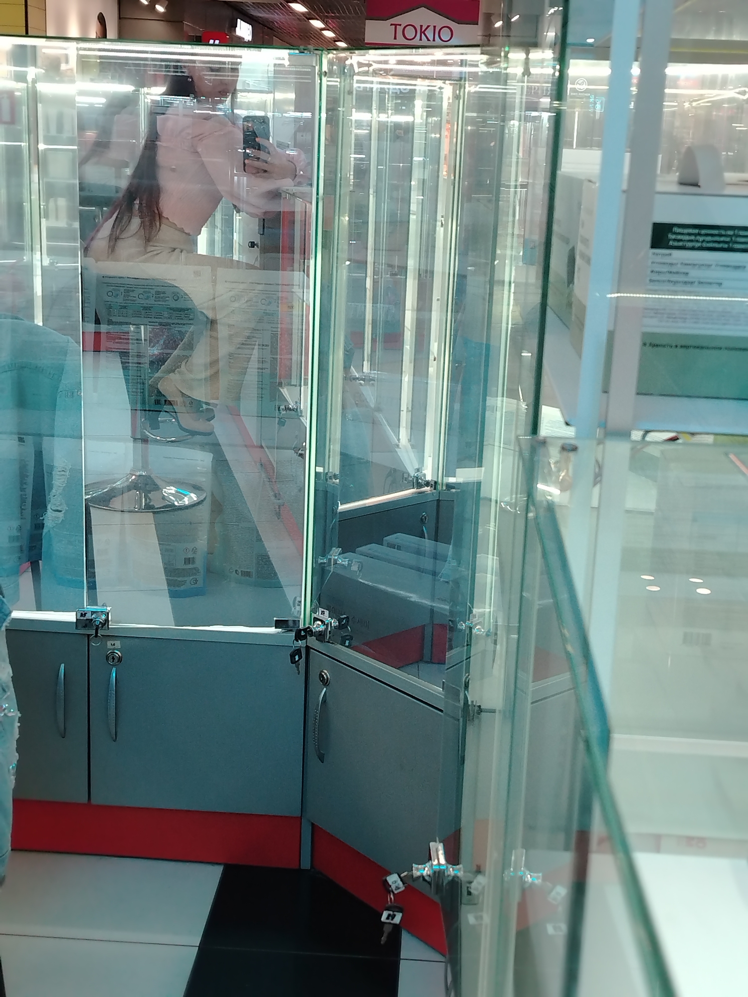 The image shows an interior view of a store, possibly a jewelry or electronics store, located in Izhevsk, Russia. A young woman is visible through a glass display case, taking a selfie with her phone. She is wearing a light pink top and beige pants. The display case is made of glass and metal, with gray cabinets and red accents at the bottom. There are several locks and keys visible on the display case doors. The store appears to be well-lit with overhead lighting. Reflections in the glass suggest a busy commercial environment. The logo "TOKIO" is visible at the top of the display case, which may indicate the name of the store or a brand.