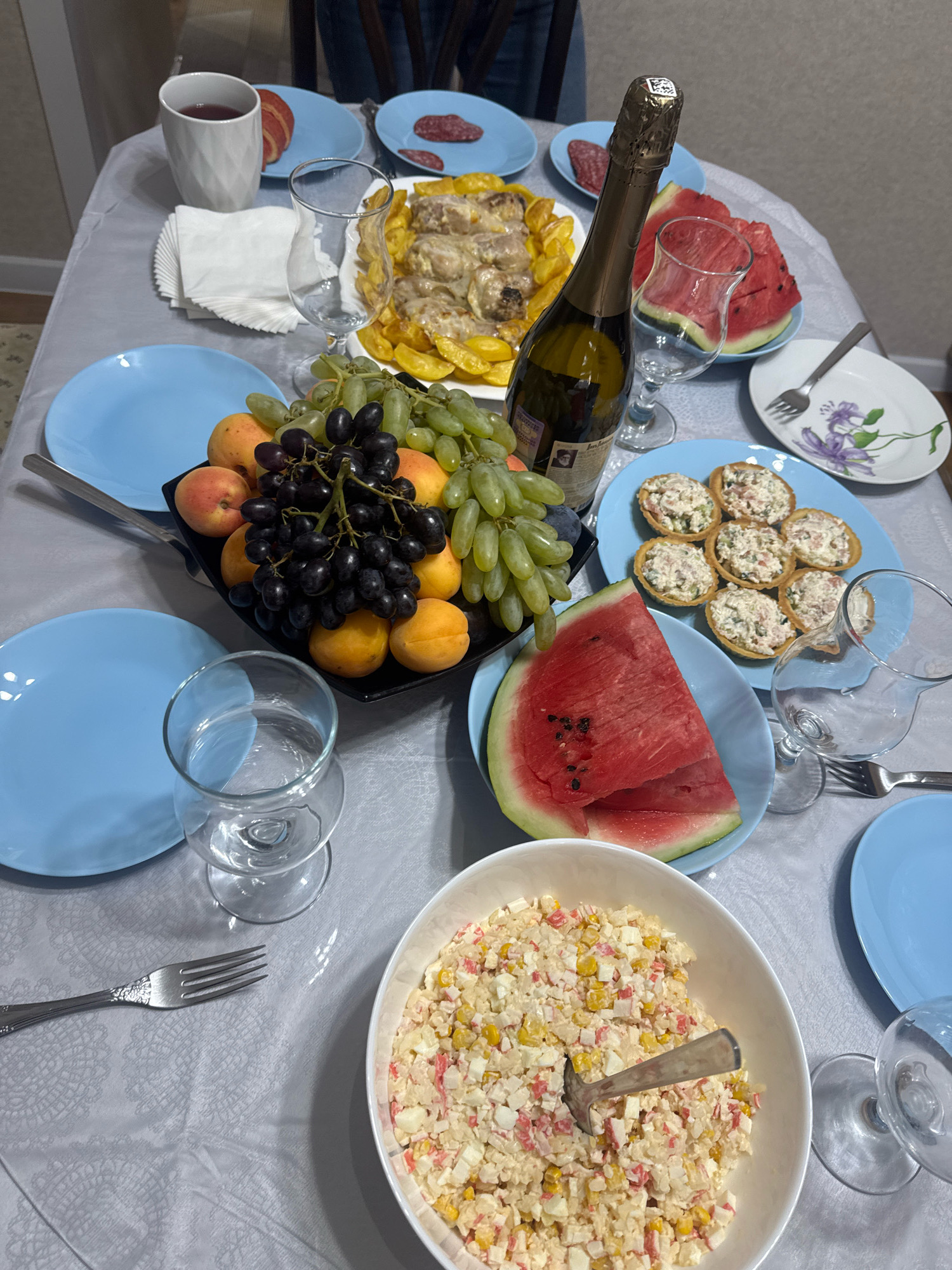 A table is set for a meal with various dishes and drinks. In the center, a bottle of champagne stands next to a sliced watermelon and a glass. To the left, a bowl of fruit, including grapes and apricots, is arranged with a fork. Further left, a white cup with red liquid and a napkin are placed next to a blue plate with sliced meat. Behind the champagne, a plate of food, possibly chicken and potatoes, is visible. In the foreground, a bowl of salad with corn and crab sticks is served with a spoon. Several blue plates and glasses are scattered around the table, indicating a gathering. The table is covered with a gray tablecloth patterned with lace-like designs. The scene appears to be an indoor dining setting. This image does not provide any information about the city of Novosibirsk, Russia.