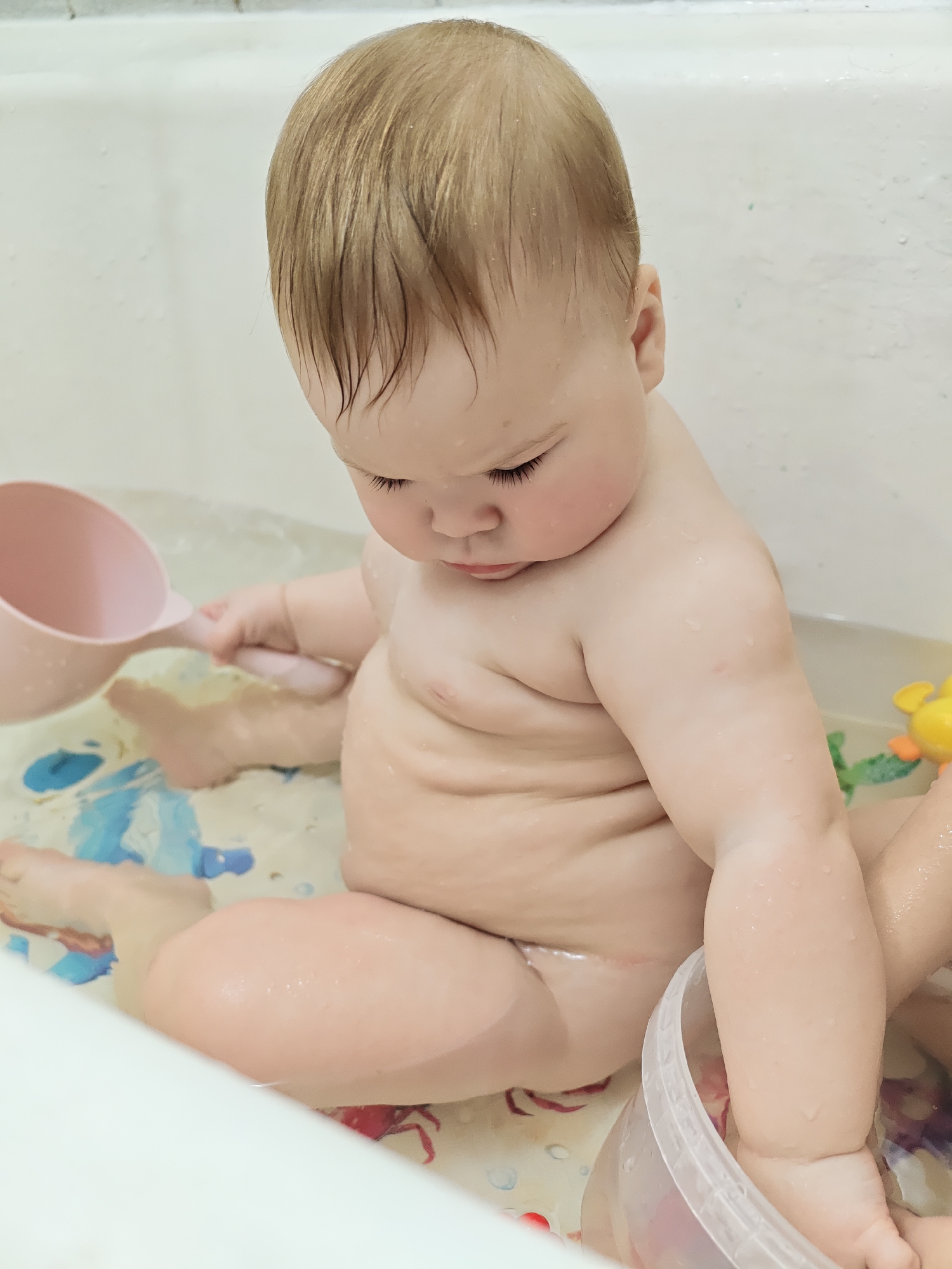 A baby sits in a bathtub with a small amount of water covering the bottom. The baby is fair-skinned with blonde hair. They sit in a seated position with legs spread, holding a pink scoop in their left hand. A clear bucket with red markings sits to their right. The bathtub has a white, shiny surface. The floor of the tub is covered with colorful, patterned anti-slip mats. Toys can be seen floating in the water. Based on the generic appearance of the bathroom and lack of specific local details, it is impossible to confirm this image was taken in Chaikovsky, Russia, or any other specific location.