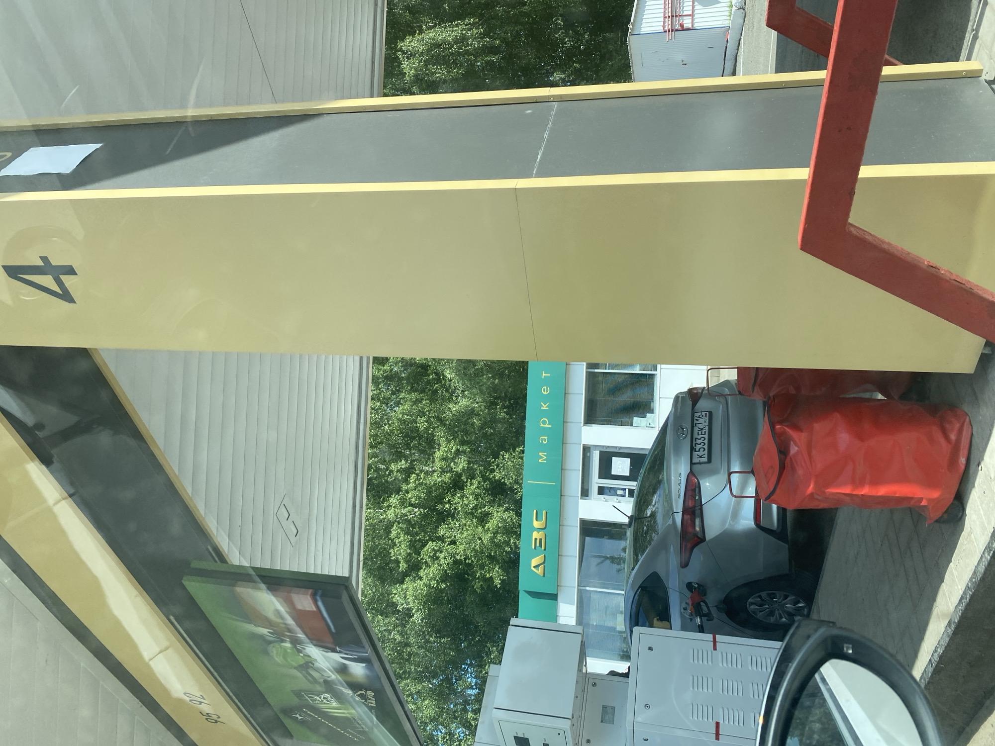 The image depicts a scene at a gas station in Vasilyevo, Russia. A silver Hyundai Solaris is parked at a fuel pump, with the fuel nozzle inserted into the car's fuel tank. The license plate of the car is visible and reads "K 533 EK 716". To the right of the car, two large red bags with wheels are situated on the ground. In the background, a building with green signage reading "Азс маркет" (which translates to "Gas Station Market") is visible. The gas station appears to be equipped with multiple fuel dispensers. A large yellow pillar with the number "4" prominently displayed on it stands to the right of the frame. The environment is outdoors with lush green trees forming the backdrop. The lighting suggests it is daytime.