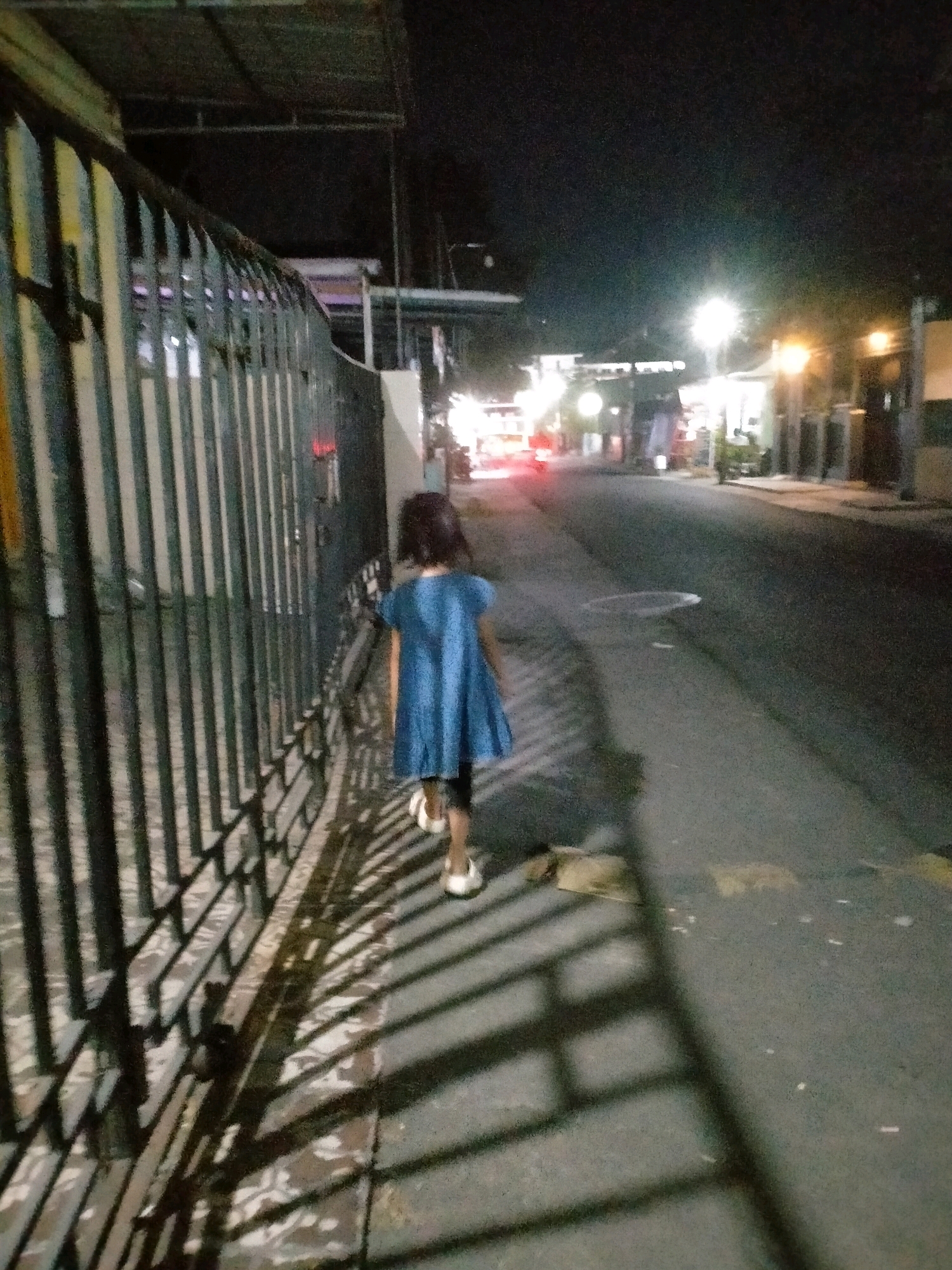 A young girl wearing a blue dress walks along a sidewalk next to a metal fence at night. The sidewalk is adjacent to a street with streetlights illuminating the scene. Buildings and trees are visible in the background. The ground is wet, suggesting it may have rained recently. This scene is located in Kasihan, Indonesia.