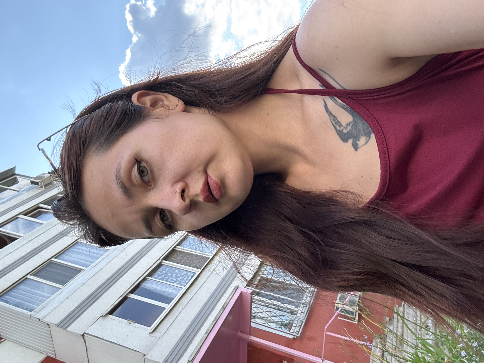 A woman with long, dark brown hair and light green eyes is pictured in a selfie outdoors. She is wearing a maroon tank top with a bird tattoo on her left chest. She has a nose ring and wears eyeliner and lipstick. The background shows a multi-story building with white and gray siding, pink accents, and windows. The sky above is blue with scattered white clouds. The location is specified as Naberezhnye Chelny, Russia.