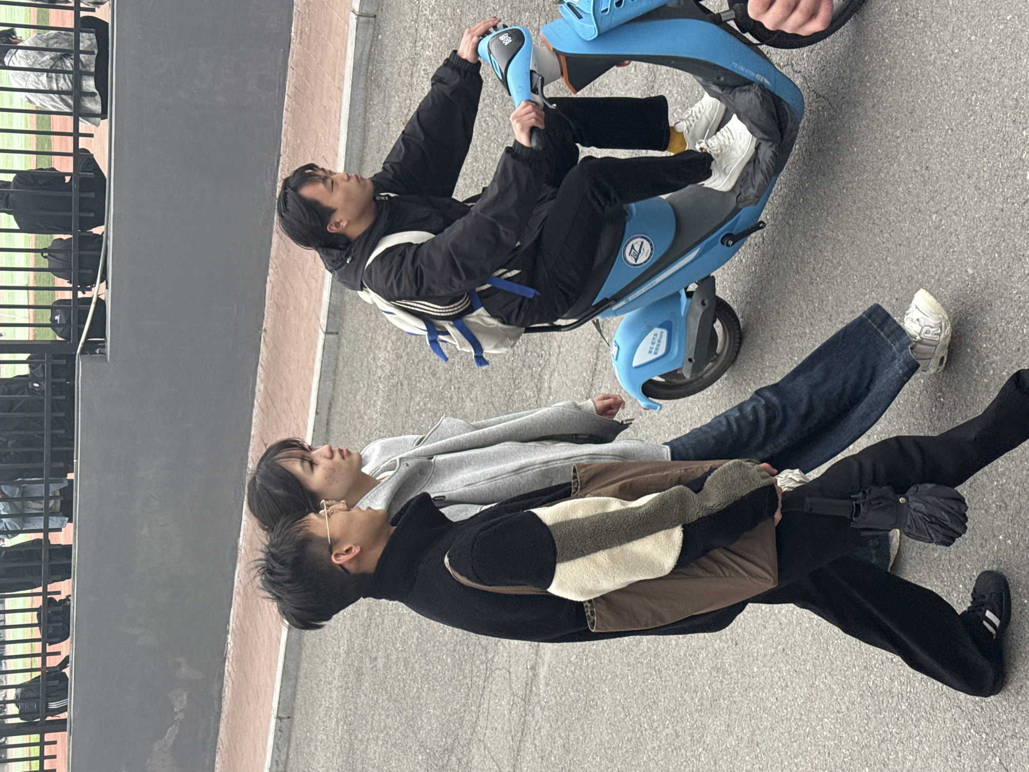 Three individuals occupy an exterior space of Zhongtun, China. One person is seated on a light blue electric scooter, wearing a black jacket, black pants, and white sneakers with yellow socks. Another person is standing, wearing a grey hoodie and jeans. The third individual is wearing a black jacket, brown jacket, black pants, and black sneakers with white stripes. An umbrella is present. A fence is on the left side of the image.