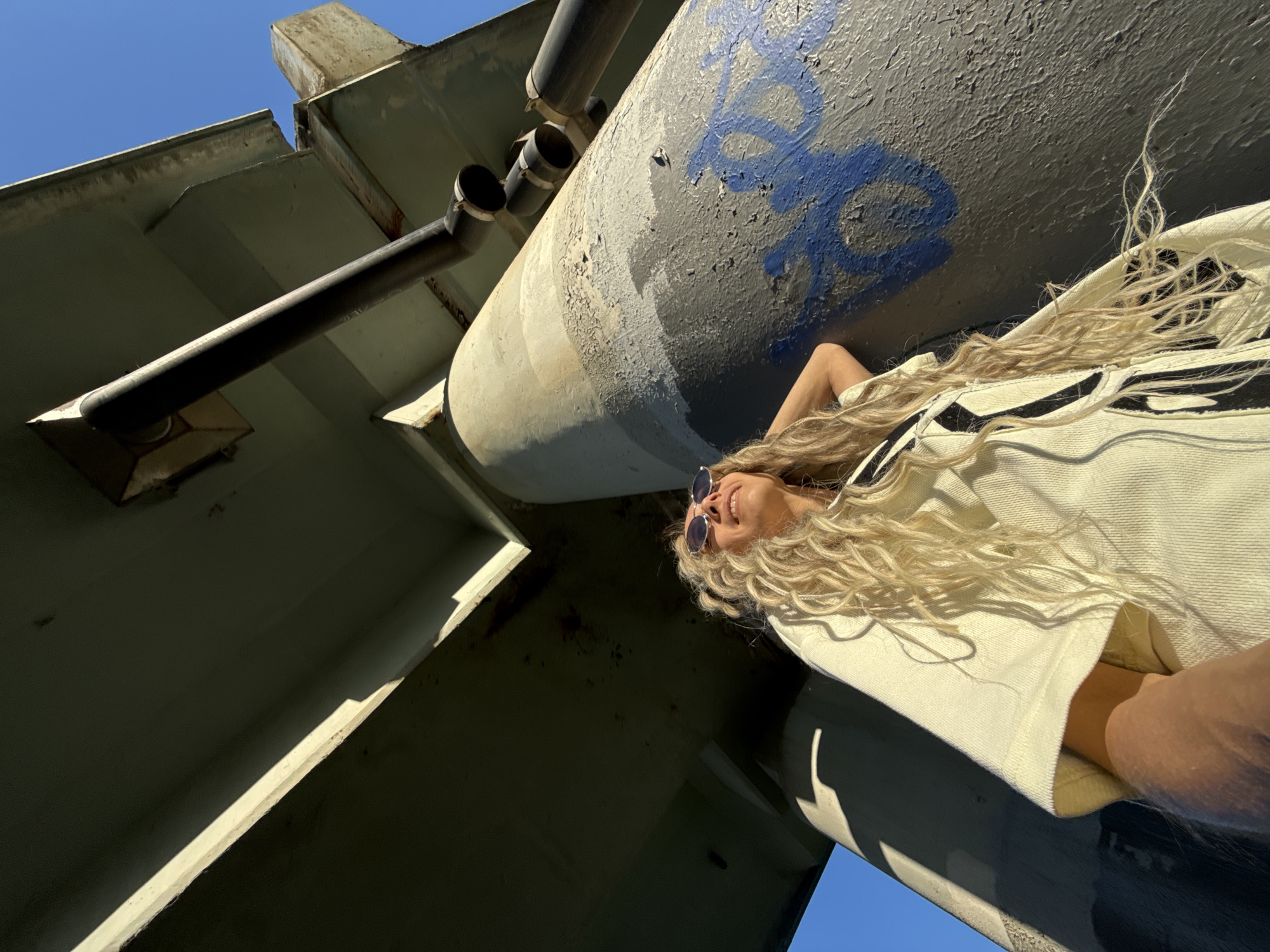 An outdoor, low-angle shot depicts a woman with long, wavy blonde hair and sunglasses, looking upwards with a slight smile, positioned under a large concrete overhead structure. She leans against a weathered concrete pillar that displays peeling grey paint and blue spray-painted graffiti. Above, the underside of a bridge or elevated roadway is visible, featuring several dark pipes attached to its surface. Intense natural sunlight illuminates the scene, creating strong shadows and highlighting the woman's light-colored, possibly distressed, garment. The background consists of a clear, bright blue sky. This scene is located in the city of Шебунино, Russia.