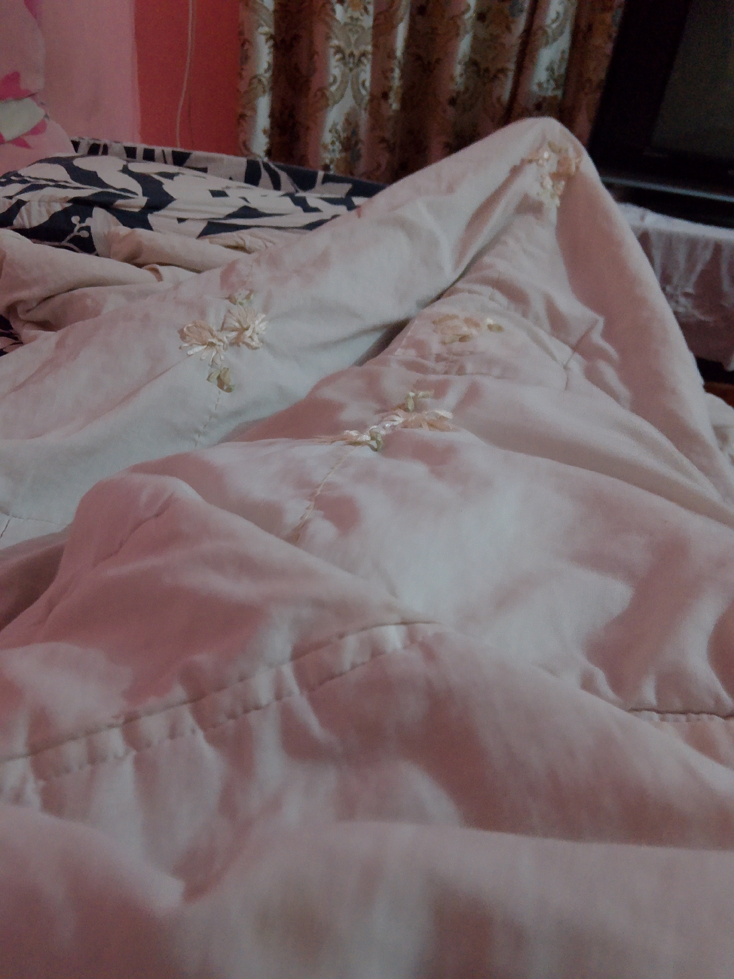 The image shows a close-up view of a rumpled white comforter with embroidered floral details. The comforter is laid out on a bed, with a patterned fabric peeking out from underneath. The background is out of focus, but appears to be a room with curtains and a wall. The lighting suggests it may be evening or a dimly lit room. There is no visible indication of the city of Mansehra, Pakistan within the image itself.