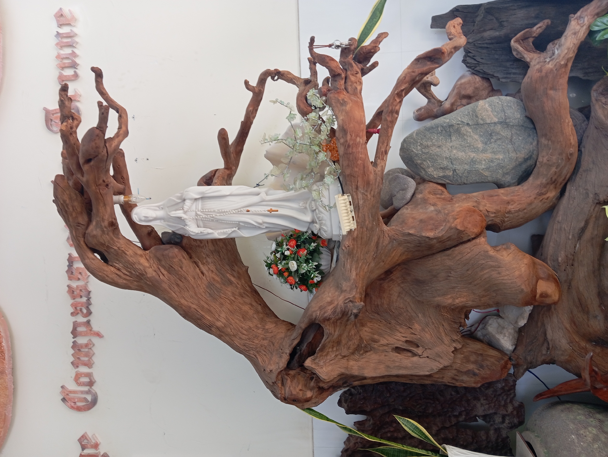 A statue of the Virgin Mary, in white, stands on a large piece of driftwood. The statue is adorned with a rosary. To the left of the statue is a small bouquet of white and orange flowers in a white vase. The driftwood is sculpted into a branching form, with various smaller pieces of wood and rocks incorporated into its base. The entire arrangement is set against a white wall that has some text written on it, partially obscured by the driftwood. The text appears to be in a decorative font and includes the words "Compass" and "Fortune". The scene is illuminated by natural light.