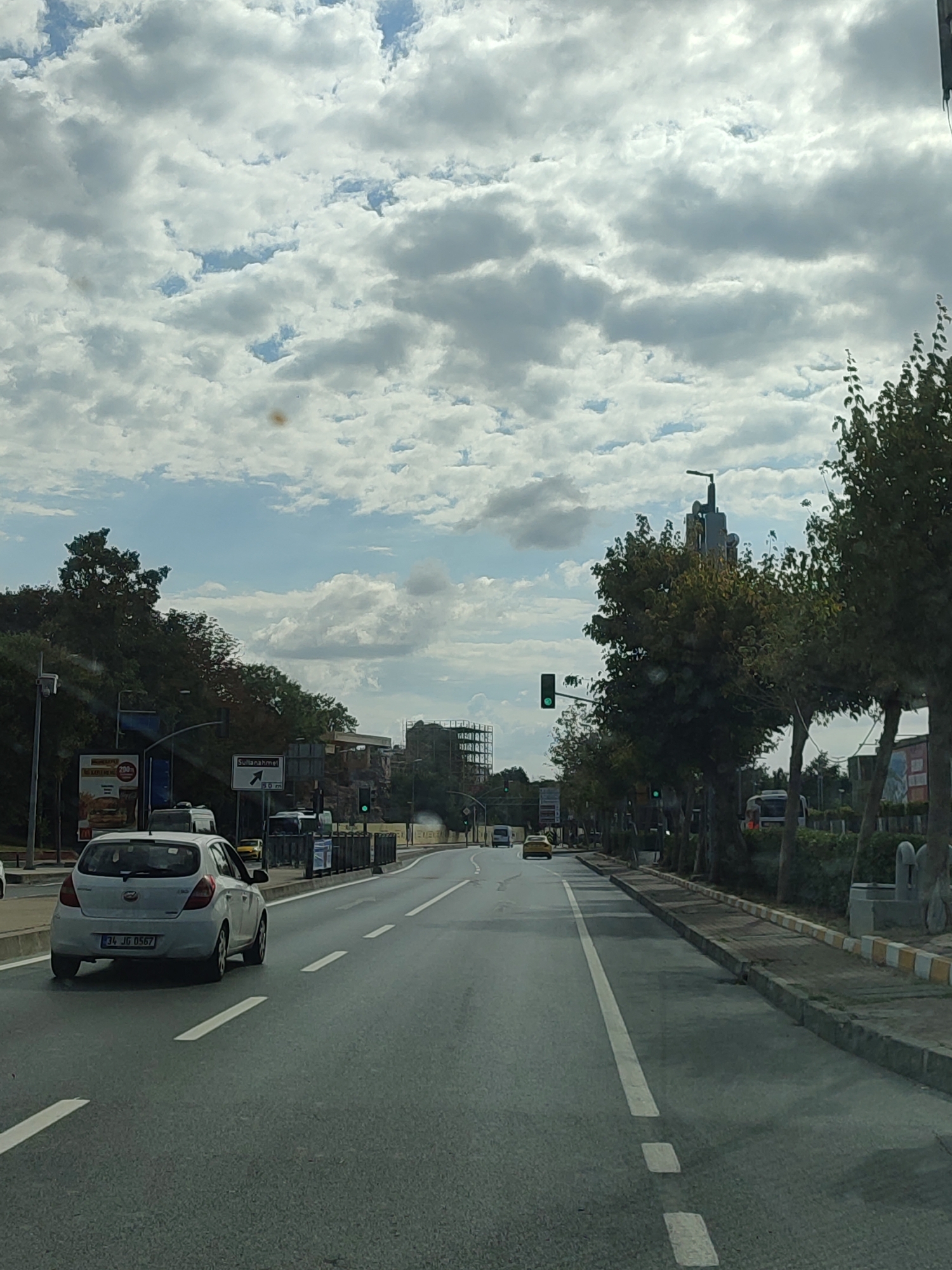 The image captures a street scene in Istanbul, Turkey, looking down a multi-lane road lined with trees and buildings. A white car with the license plate "34 JG 0567" is in the foreground, on the right side of the road. Traffic lights are visible, one showing green. A sign points towards "Sultanahmet" with a distance of "50 m". The sky is partly cloudy, with scattered white and grey clouds against a blue sky. Trees line the right side of the road, and construction is visible in the distance on the left. The road surface is asphalt, with white lane markings.