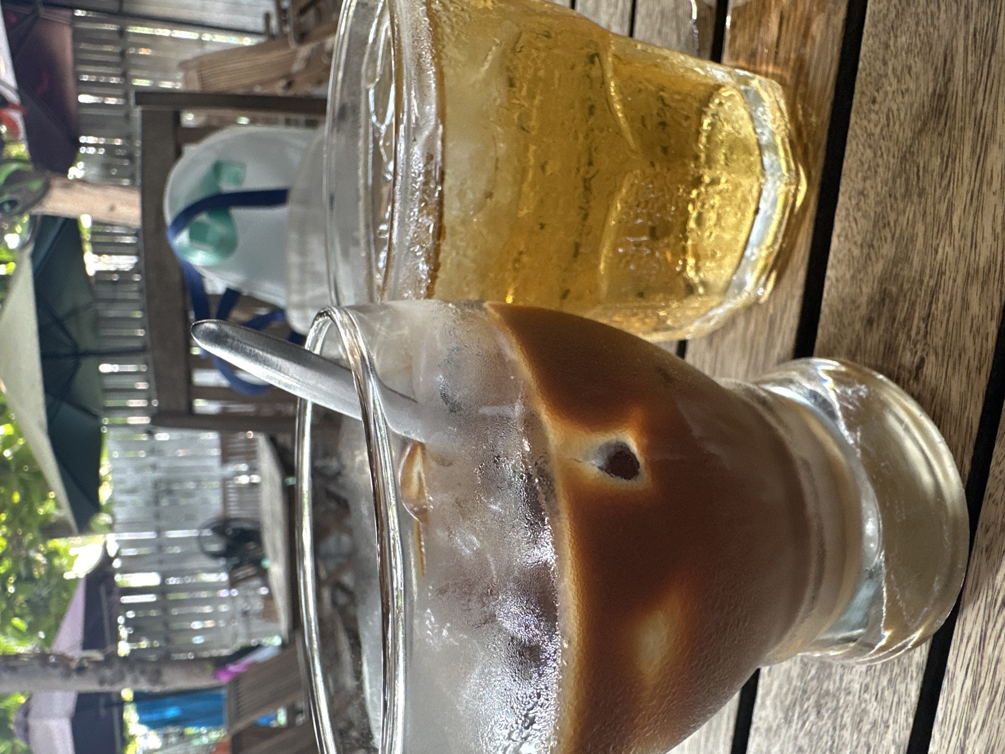 The image shows two drinks in clear glasses on a wooden surface. The drink in the foreground has a layered appearance, with a dark brown liquid on top and a lighter, milky substance beneath. A metal spoon is partially submerged in the drink. The glass is visibly frosted, suggesting it contains ice. Behind it, another glass is filled with a golden-colored liquid, likely iced tea, also with visible condensation. The drinks are likely on a table at a cafe or restaurant. In the blurred background, there's a partial view of an outdoor setting with a woven fence, glimpses of greenery, and a partial view of what looks like a folded white fabric and some additional objects. Based on the provided information, the location is Nha Trang Ward, Vietnam.
