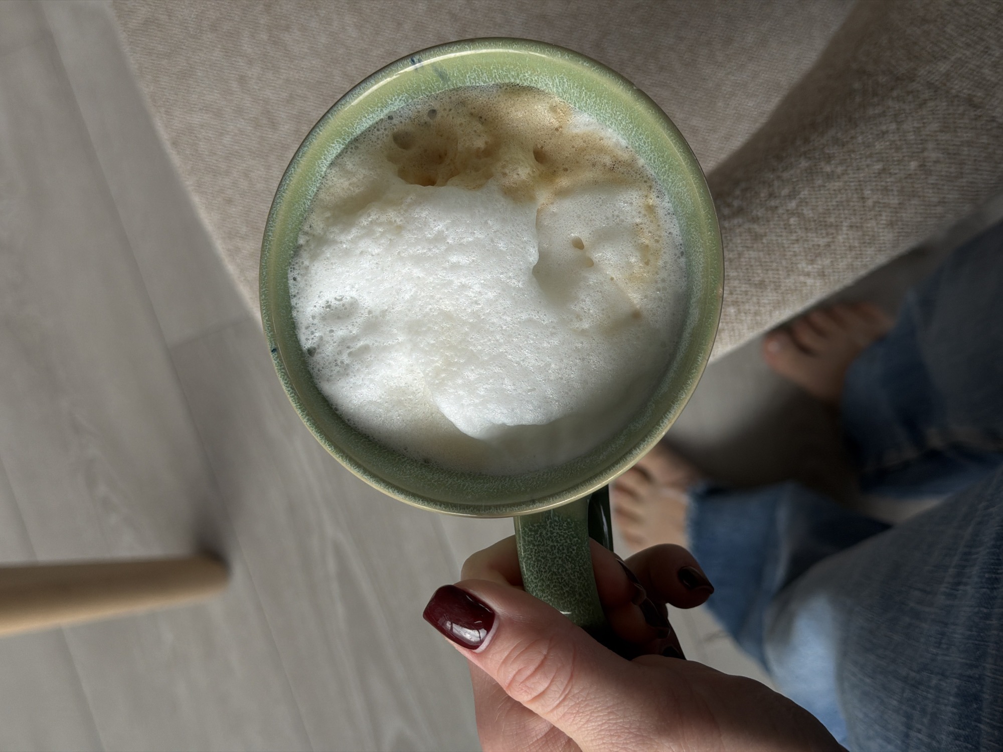 A person is holding a green mug filled with foamy coffee. The person is wearing blue jeans and has dark nail polish. Part of a wooden chair leg is visible on the left. Bare feet are visible in the background, suggesting the person is indoors. The location in Dreiliņi, Latvia, cannot be ascertained from the visual information.