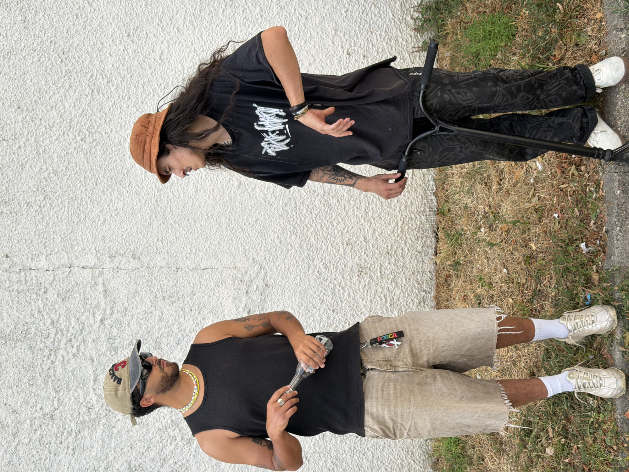 The media file displays two individuals outdoors, against a textured white wall and a grassy embankment. The upper individual, oriented towards the viewer's right, wears a black t-shirt with a white graphic design, black pants with a patterned print, and white shoes. They have long, dark hair and are wearing a brown corduroy bucket hat. Their stance suggests they are interacting with a black scooter. The lower individual, facing the viewer's left, is dressed in a black tank top, khaki shorts with frayed hems, and white sneakers with white socks. They are wearing sunglasses, a beaded necklace, and a ring on their left hand. This person is holding a clear glass bottle. A keychain with the text "POSCA" is attached to their shorts. The lighting suggests it is daytime, and the dry grass indicates warm weather. The overall scene appears to be casual and urban, possibly a street or sidewalk area in Caluire-et-Cuire, France.