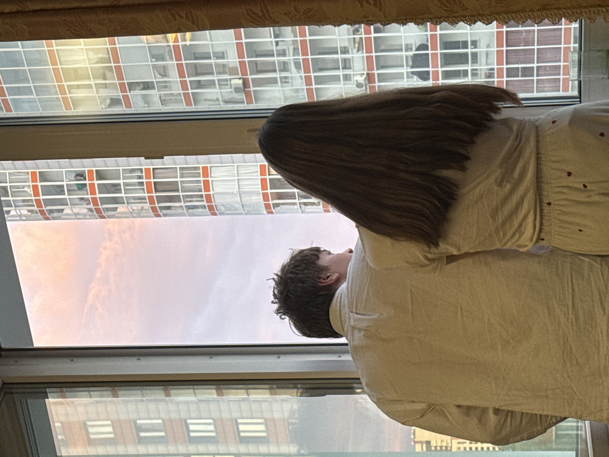 A couple looks out of a window. The location is Krasnoyarsk, Russia, as seen from the buildings outside the window. One person has long brown hair and is wearing a light-colored shirt with hearts. The second person has short dark hair and is wearing a light-colored shirt. The sky outside the window is pinkish. The window has a tan curtain above it.