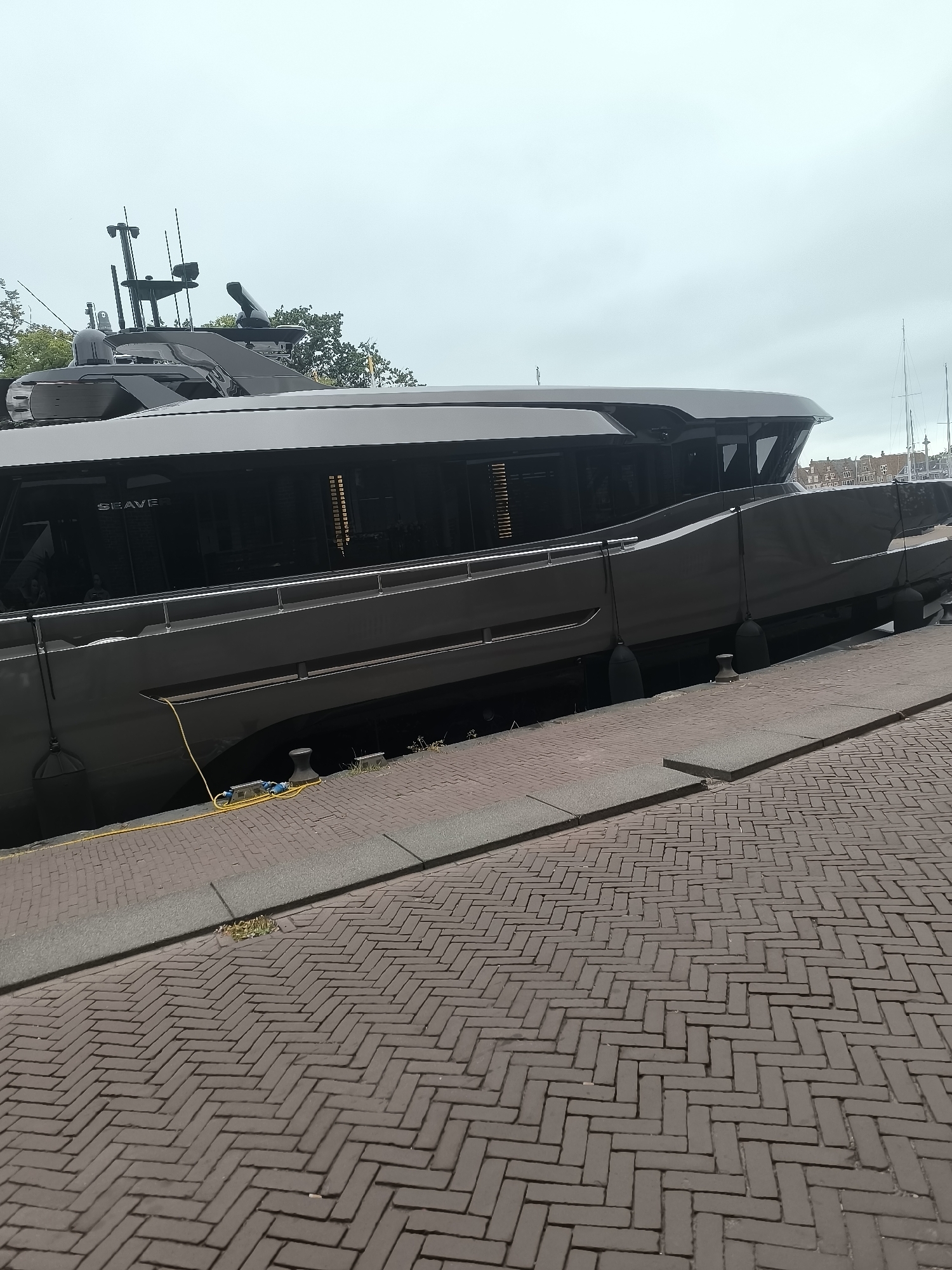 A dark gray yacht, named "SEAVERS", is docked along a paved walkway in the city of Hoorn, Netherlands. The yacht's sleek, modern design features large tinted windows and a complex array of antennas and equipment on its upper deck. A yellow electrical cable runs from the yacht to the dock, connecting to what appears to be a charging station or power source. The walkway is constructed of dark reddish-brown brick pavers arranged in a herringbone pattern. Along the edge of the walkway, dark fender buoys are secured to the yacht. In the background, other docked boats and buildings with red roofs are visible, suggesting a harbor or marina setting. The sky is overcast and a light gray, indicating a cloudy day. No people are visible in the image.