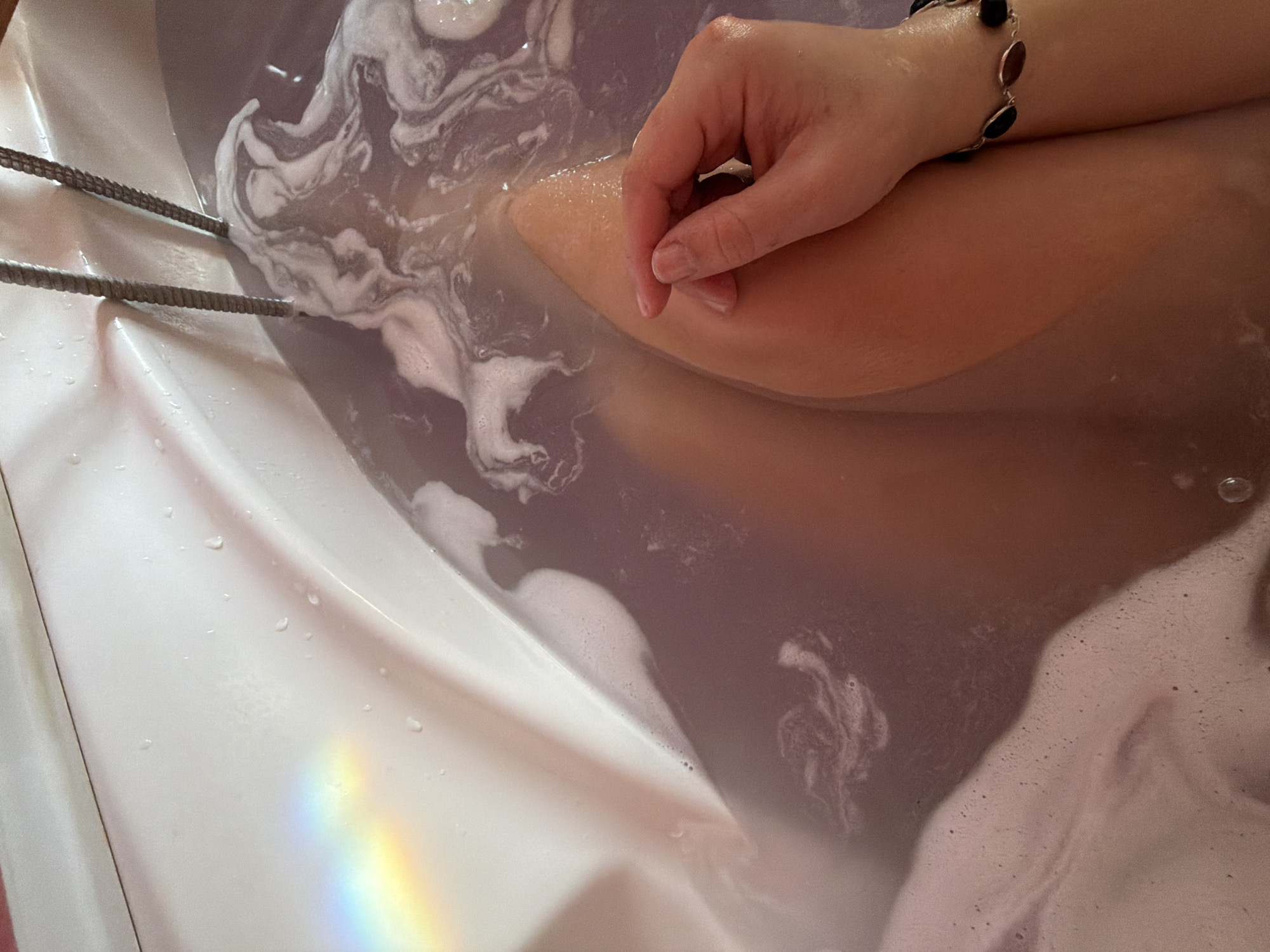 The image depicts a partial view of a person in a bathtub, likely indoors. The tub is filled with water and bath foam, exhibiting a marbleized swirl pattern. A hand with a bracelet rests on a leg submerged in the water. Two gray, braided cords are visible along the left edge of the tub. A rainbow-like reflection is present on the white tub surface. The location is assumed to be in a residence in the city of Большая Шильна, Russia.