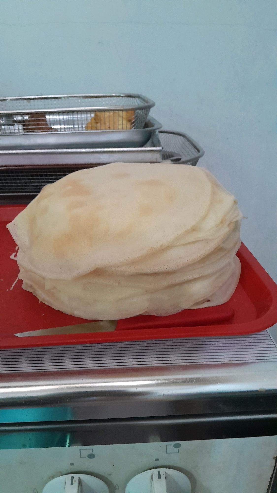 A stack of golden-brown crepes rests on a bright red plastic tray. The crepes, varying slightly in color and texture, are piled high, creating a circular shape. A thin, silver knife is placed horizontally across the front of the tray, partially obscured by the stack of crepes. In the background, two metal wire baskets are stacked, one on top of the other, containing what appears to be fried food, possibly fries. The baskets are positioned on a stainless steel surface, likely a kitchen counter. Behind the baskets, a light blue wall serves as the backdrop. The scene appears to be set in a kitchen environment, possibly in a commercial establishment or a home kitchen. The lighting is moderate, casting soft shadows. There are no people visible in the image. The image composition focuses on the crepes as the primary subject.