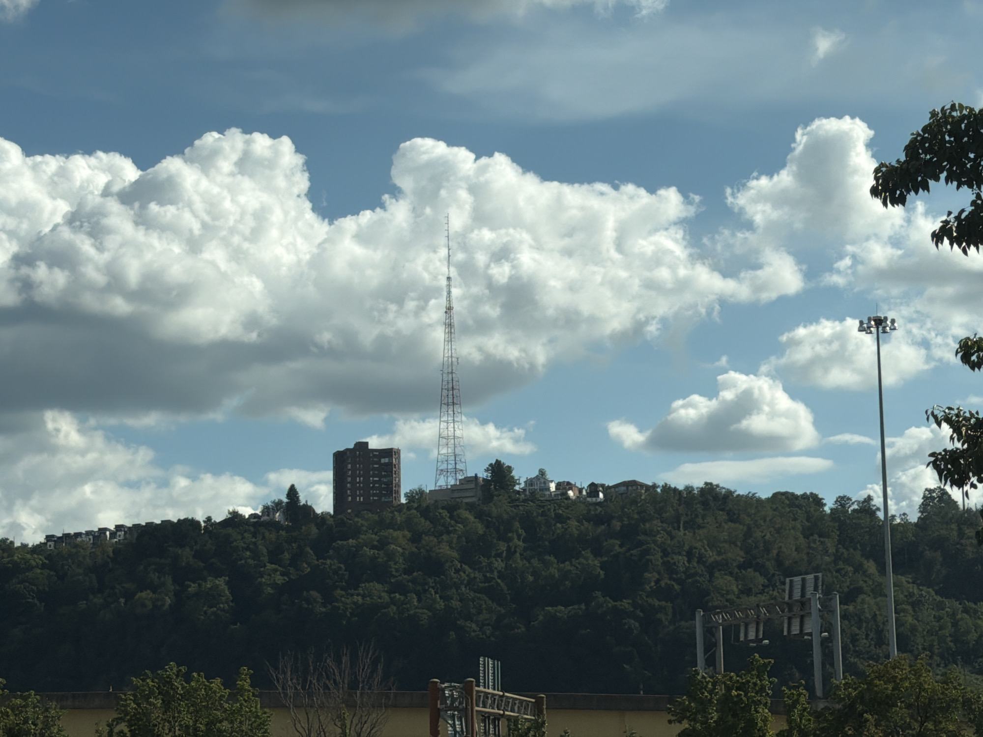 A wide shot captures a lush, tree-covered hill under a bright blue sky dotted with large, fluffy white clouds. On the crest of the hill, several buildings are visible, including a tall, dark brick building on the left and a slender radio tower in the center. A few smaller houses are scattered further along the hilltop. In the foreground, a section of a highway overpass with overhead signs is partially visible, and trees line the bottom edge of the frame. The scene appears to be a suburban or semi-urban landscape overlooking a densely wooded area.