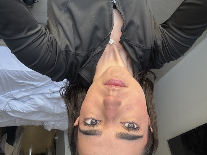 An adult female is depicted in a chest-up self-portrait, looking directly at the camera with a neutral expression. She has dark brown hair, prominent dark eyebrows, and dark brown eyes with visible mascara. Her complexion shows redness on the cheeks and nose. She is wearing a dark-colored, likely black or dark grey, full-zip athletic jacket, partially unzipped. A thin silver-colored chain necklace with a circular, coin-like pendant is visible at her neck. The background on the left features a section of a dark flat-screen television mounted on a light-colored wall. The background on the right shows an unmade bed with white bedding. Beyond the bed, a brightly lit interior space, possibly a hallway or adjacent room, is visible. Strong natural light illuminates the scene from the left. The setting is indoors, consistent with a private residence or hotel room located in the city of Unknown, Unknown.