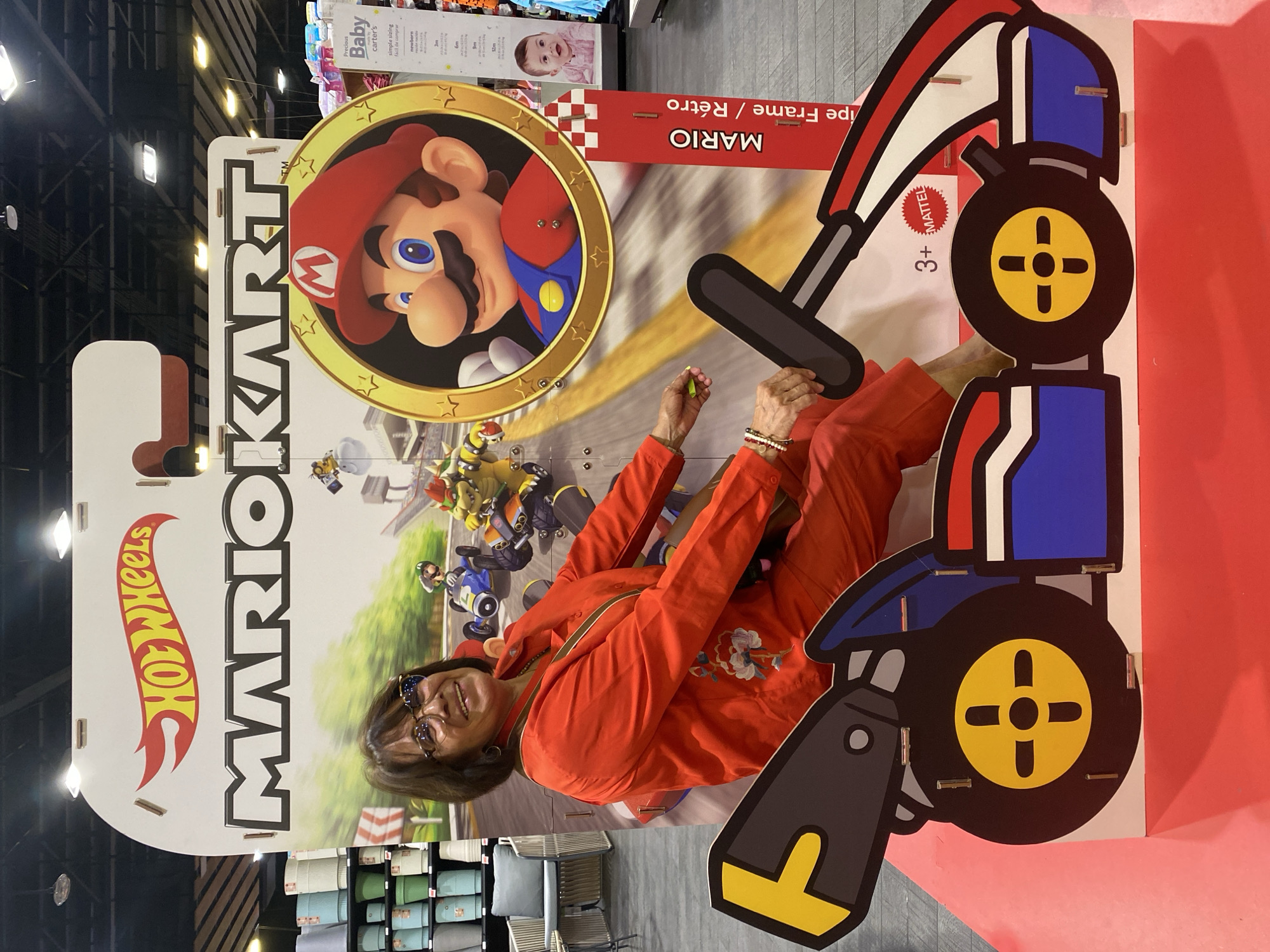 A woman in an orange outfit poses with a life-sized, stand-up MarioKart advertisement. The woman is seated within the frame of the advertisement, giving the impression that she is driving the MarioKart. The sign is positioned indoors, with retail items and store fixtures in the background. The sign advertises "MarioKart" in large letters alongside the "Hot Wheels" logo. It also features an image of Mario and other characters, as well as cartoon-style illustrations of a MarioKart. It also says "3+ MATTEL MARIO ipe Frame / Retro Baby carters new born." The location appears to be a retail store. This image does not provide enough information to determine the specific location in San Bernardino Tlaxcalancingo, Mexico.
