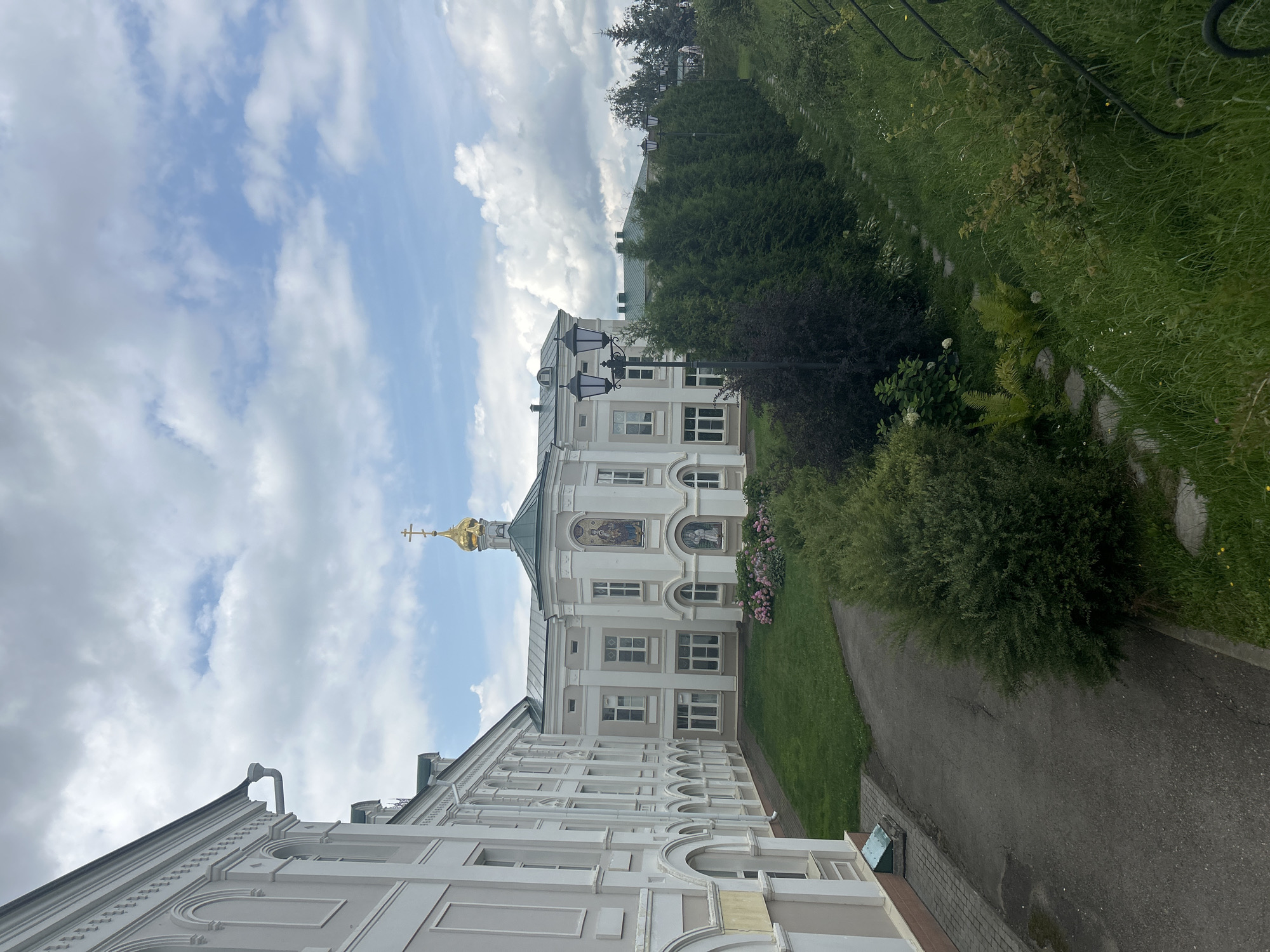 The image shows a view of the Seraphim-Diveyevo Monastery in Diveyevo, Russia. The monastery's main building, painted a pale cream color with white trim, features a green roof topped with a golden dome and cross. The facade includes arched windows and a mosaic depicting religious figures. Adjacent to the main building is a long, white structure with arched openings, suggesting a cloister or residential wing. A paved pathway curves through a grassy courtyard in front of the buildings. Landscaping includes manicured green lawns, a line of tall, dense evergreen trees, and various bushes and flowering plants, including hydrangeas. Several ornate black lampposts are visible along the pathway. The sky is partly cloudy with white cumulus clouds against a blue backdrop. The scene appears peaceful and well-maintained, reflecting the spiritual nature of the location.